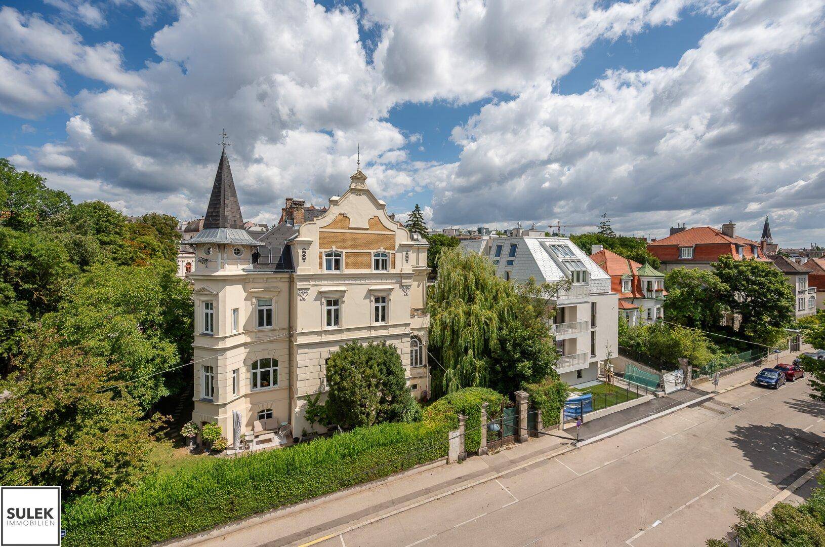 Historisches Gebäude mit markantem Turm neben einem modernen Neubau in einer grünen Umgebung.