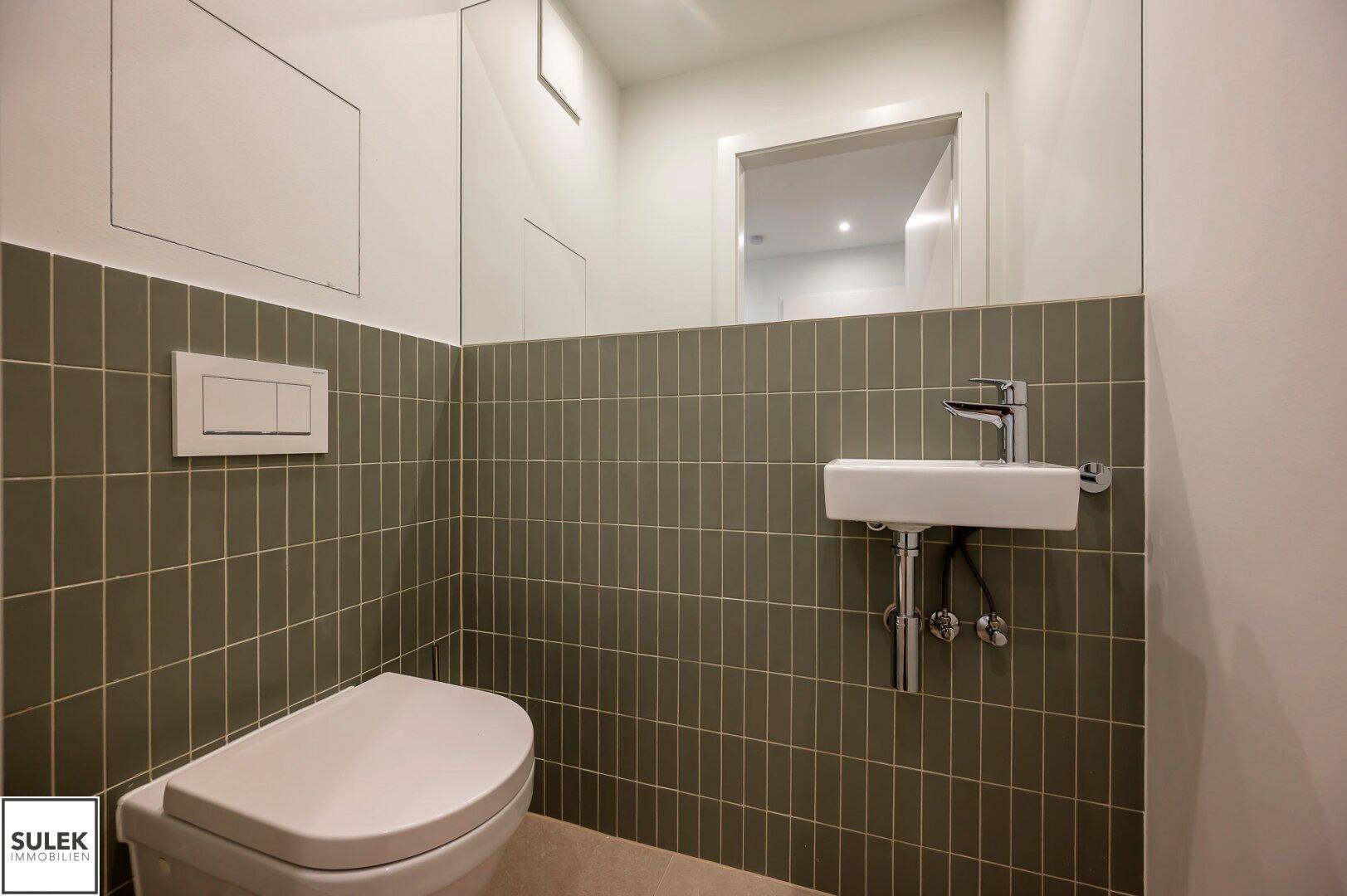 Gäste-WC mit Wand-WC, kleinem Waschbecken und grünen Fliesen an der Wand.