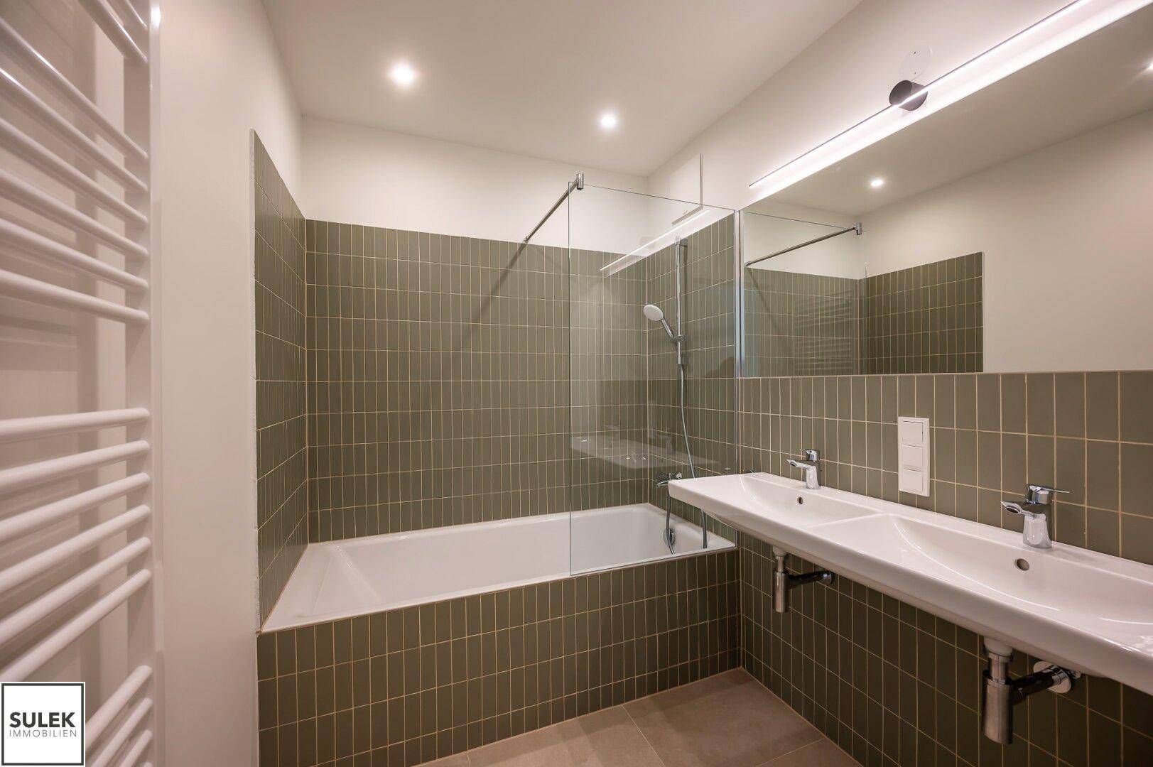 Modernes Badezimmer mit Badewanne, Doppelwaschbecken und grünen Wandfliesen.