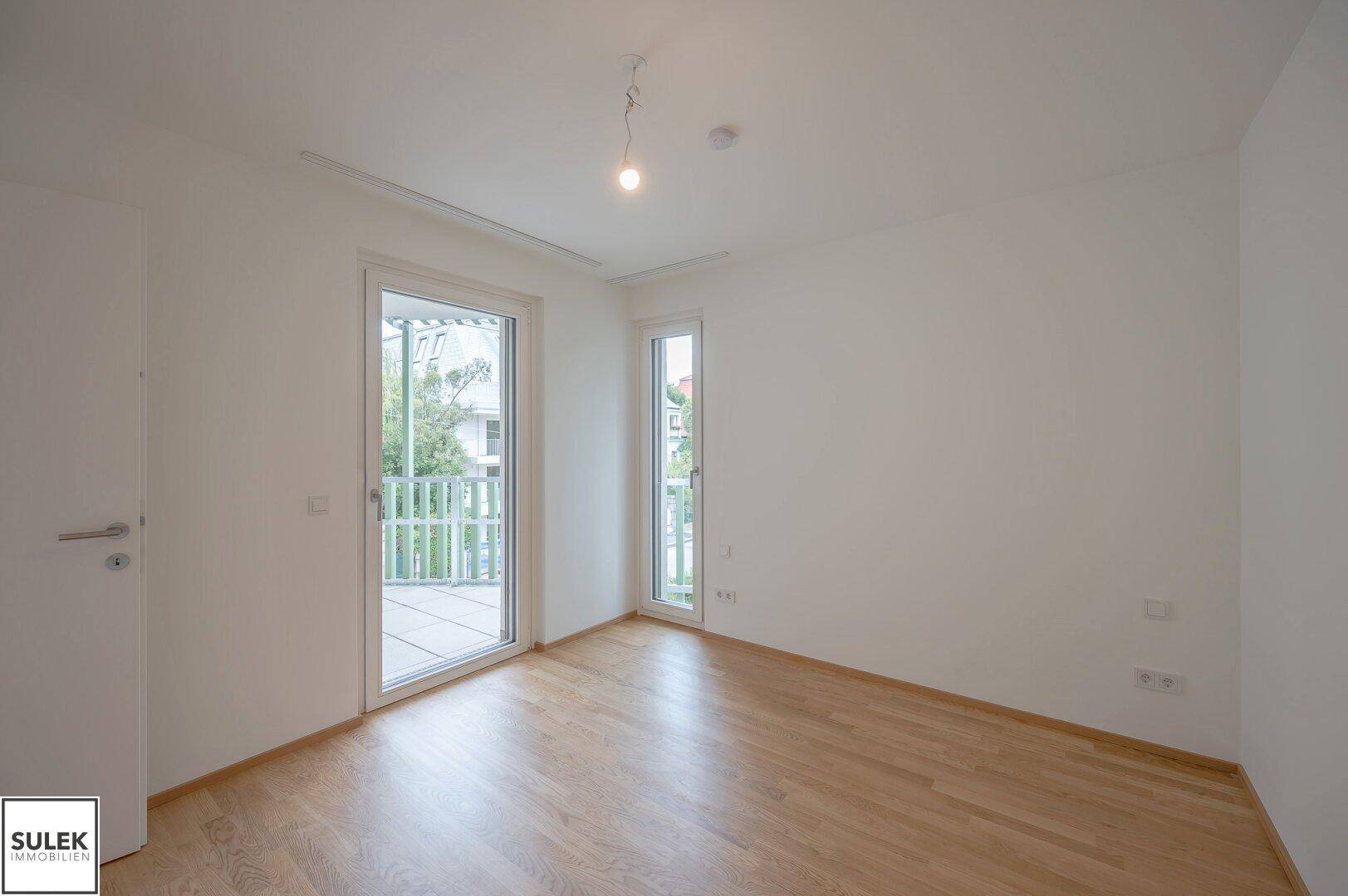 Leeres Schlafzimmer mit Parkettboden, großen Fenstern und Balkonzugang.