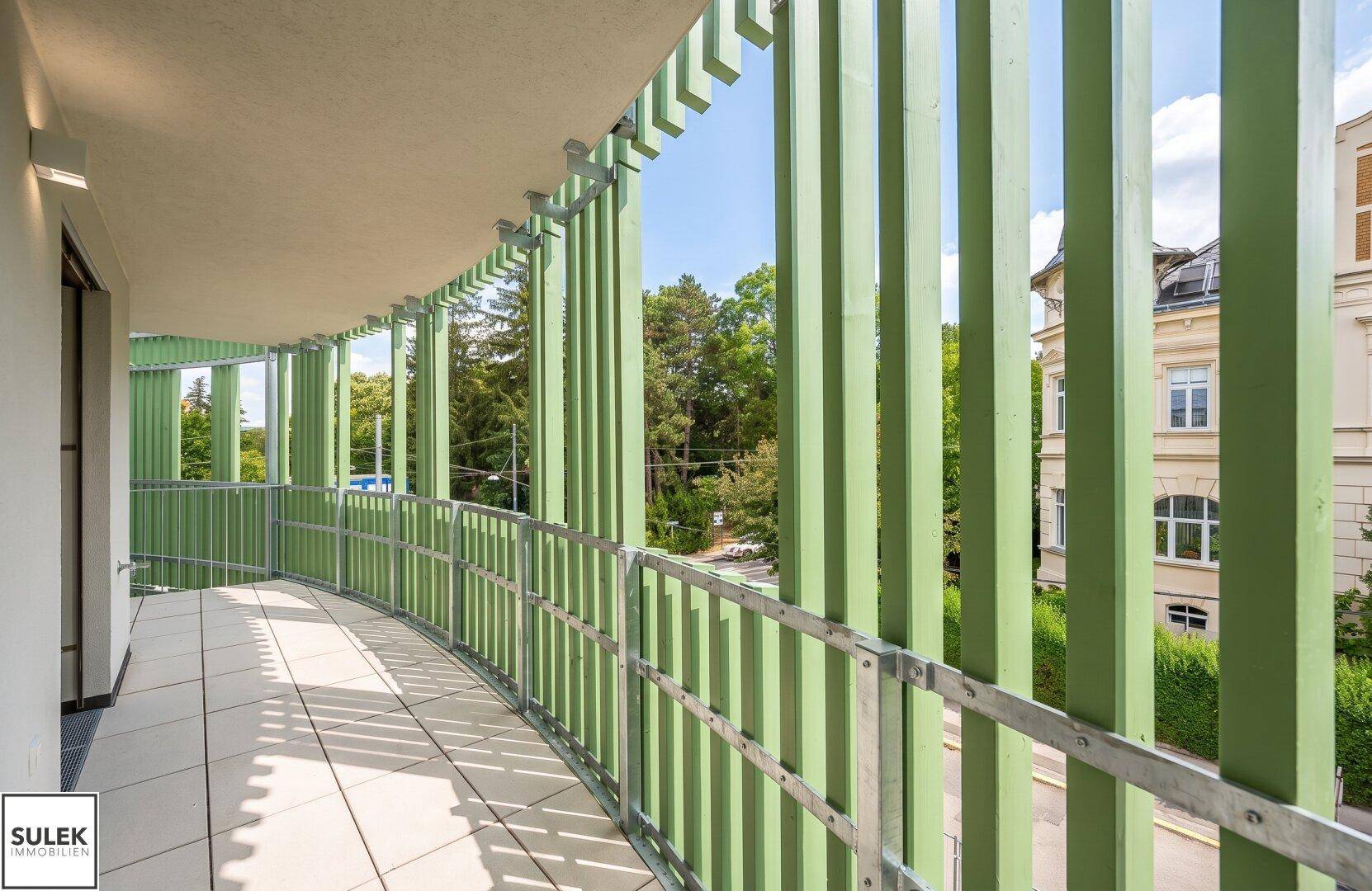 Langer Balkon mit grünen Lamellen und Fliesenboden, umgeben von Bäumen.
