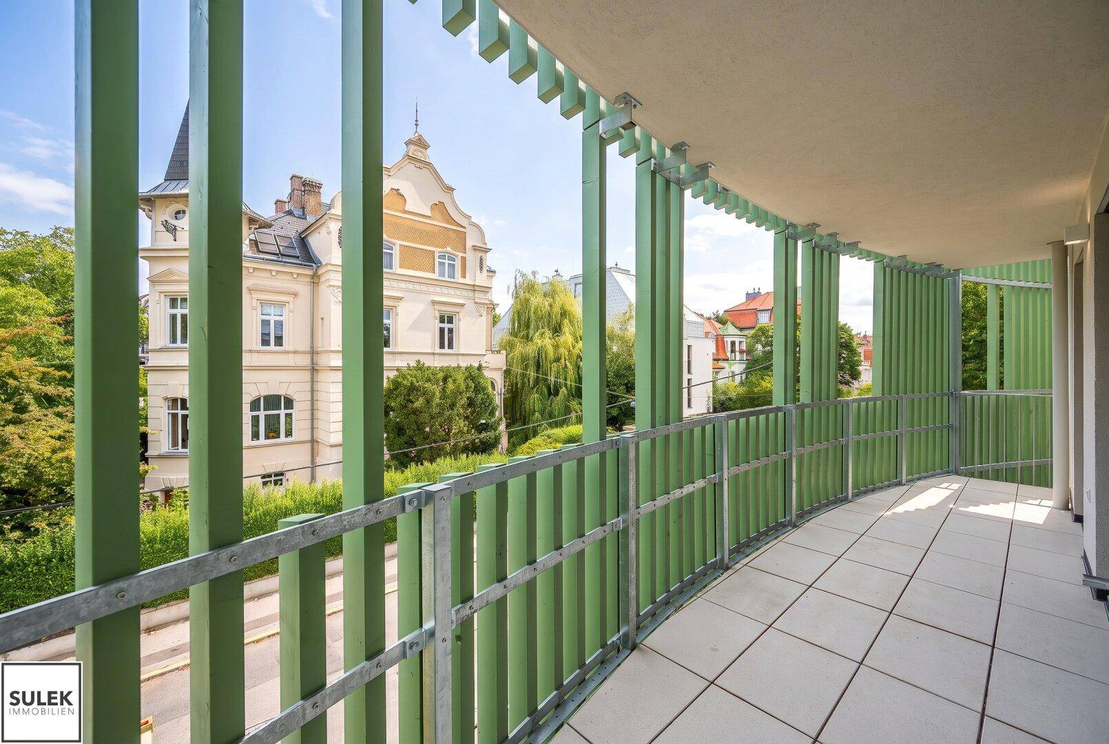 Balkon mit grünen Lamellen und Fliesenboden, bietet einen Blick auf ein historisches Gebäude.
