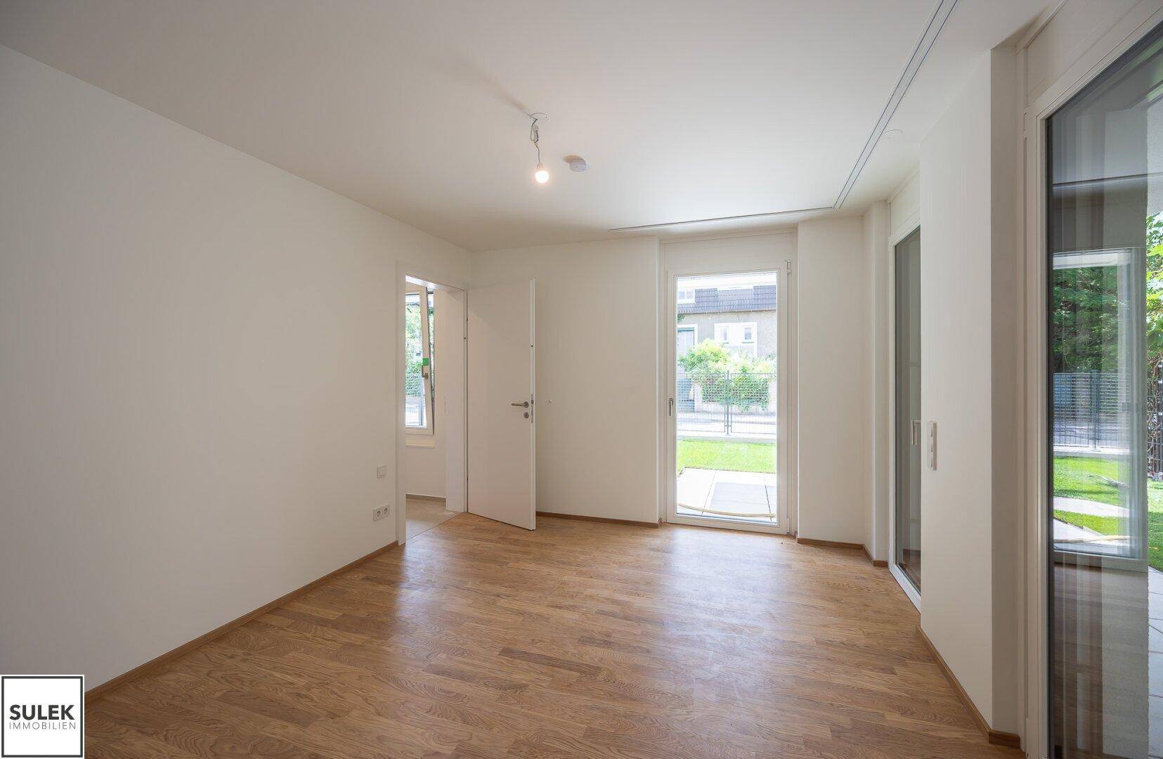 Heller, leerer Raum mit Holzboden und großen Fenstertüren zum Gartenbereich.