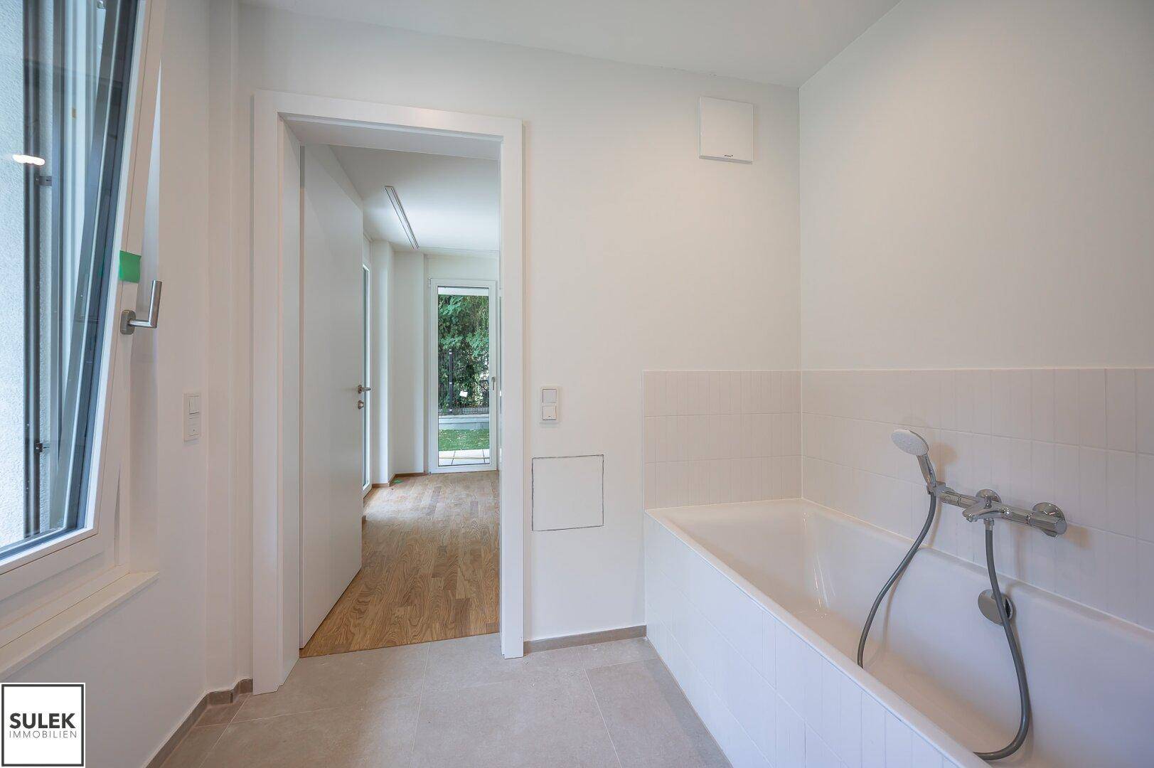 Helles Badezimmer mit Badewanne und Fenster, das den Blick in den Flur freigibt.