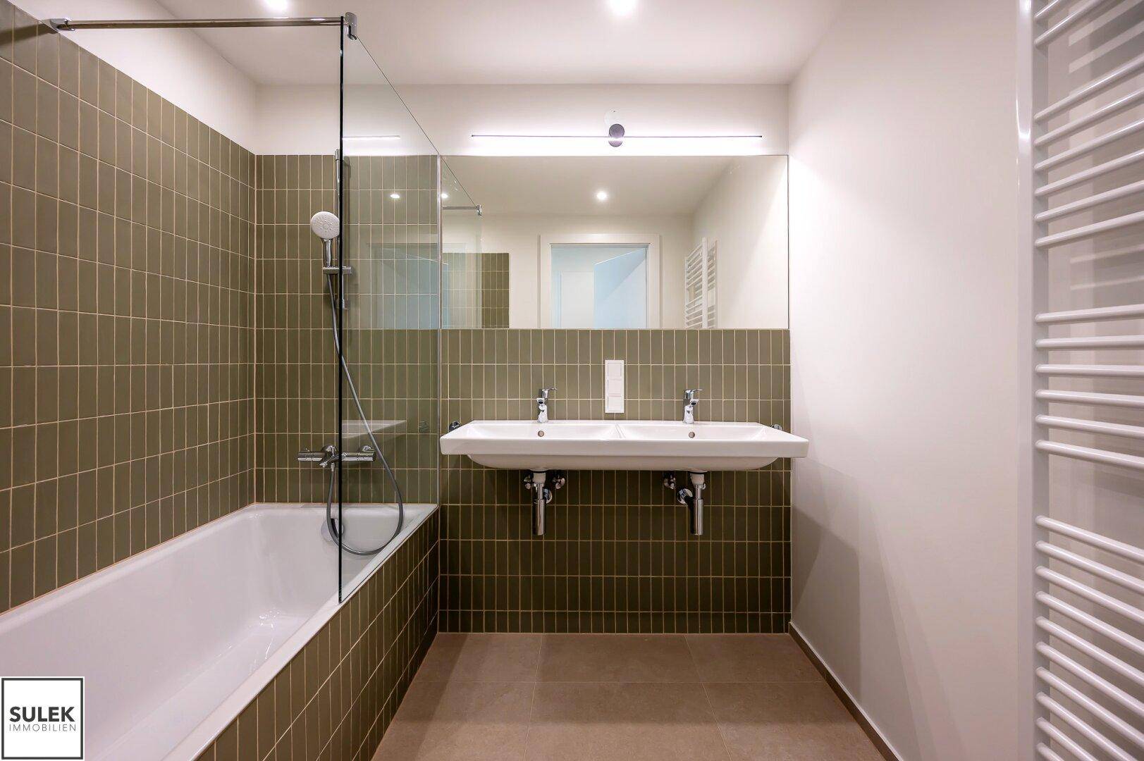 Modernes Badezimmer mit Badewanne, Doppelwaschbecken und grünen Wandfliesen, ausgestattet mit Handtuchheizkörper.