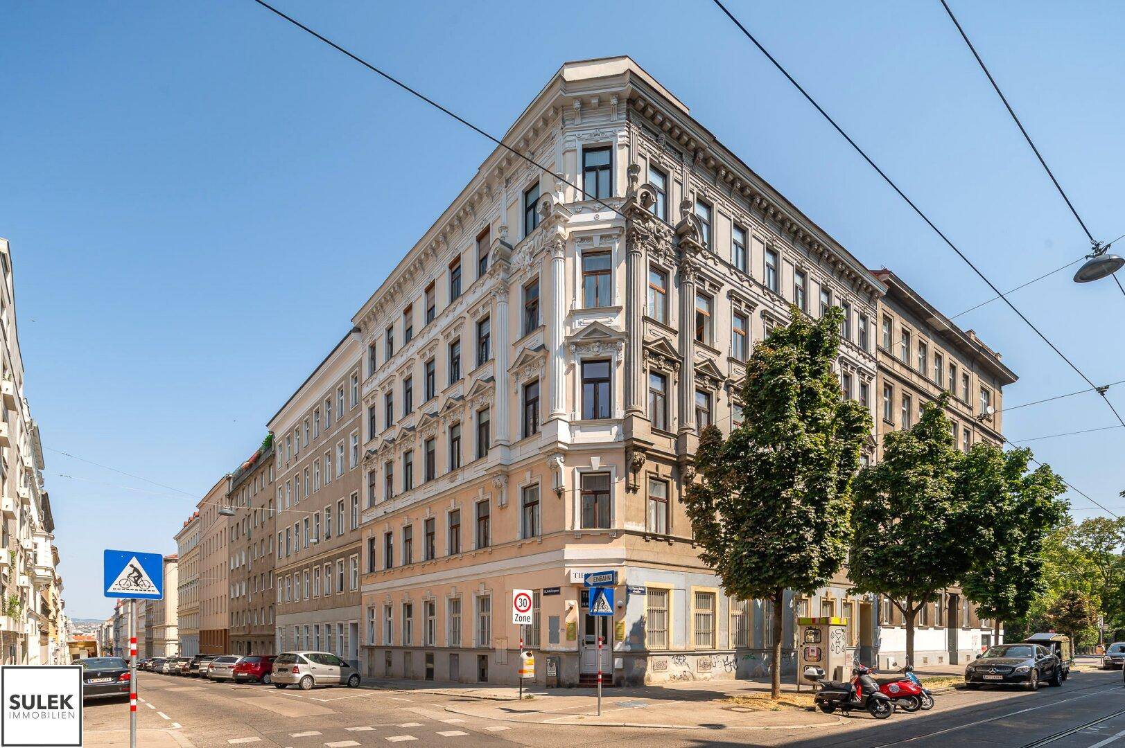 Eckgebäude in einer städtischen Umgebung mit klassischer Architektur und Bäumen.