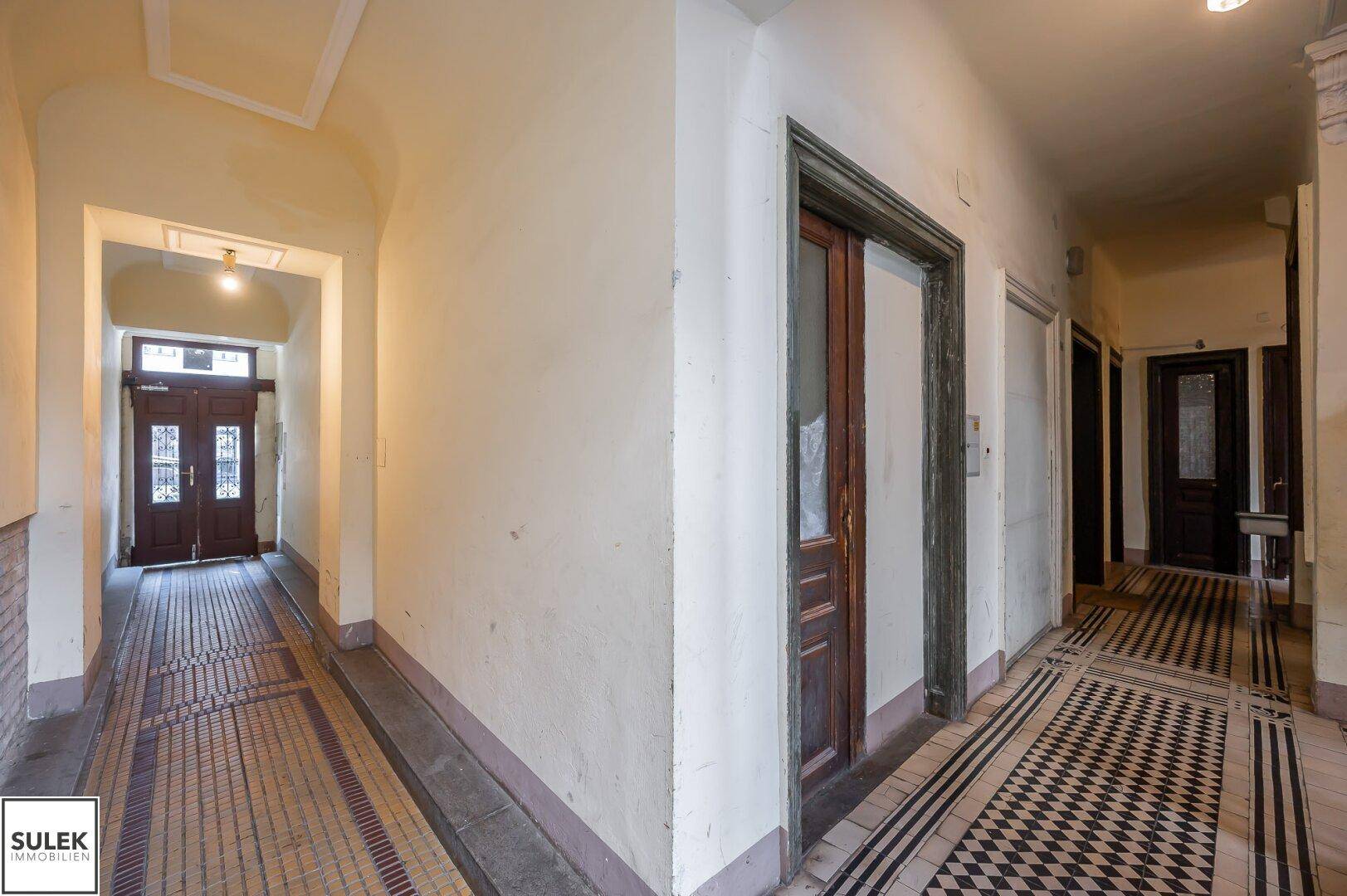 Langer, heller Flur in einem Altbau mit gemustertem Fliesenboden und Holztüren.