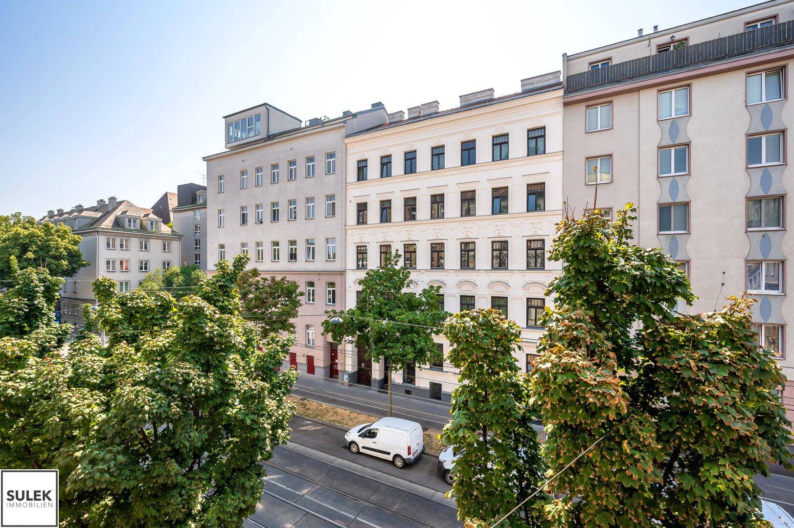 Blick auf eine belebte Straße mit Bäumen und historischen Wohngebäuden unter blauem Himmel.