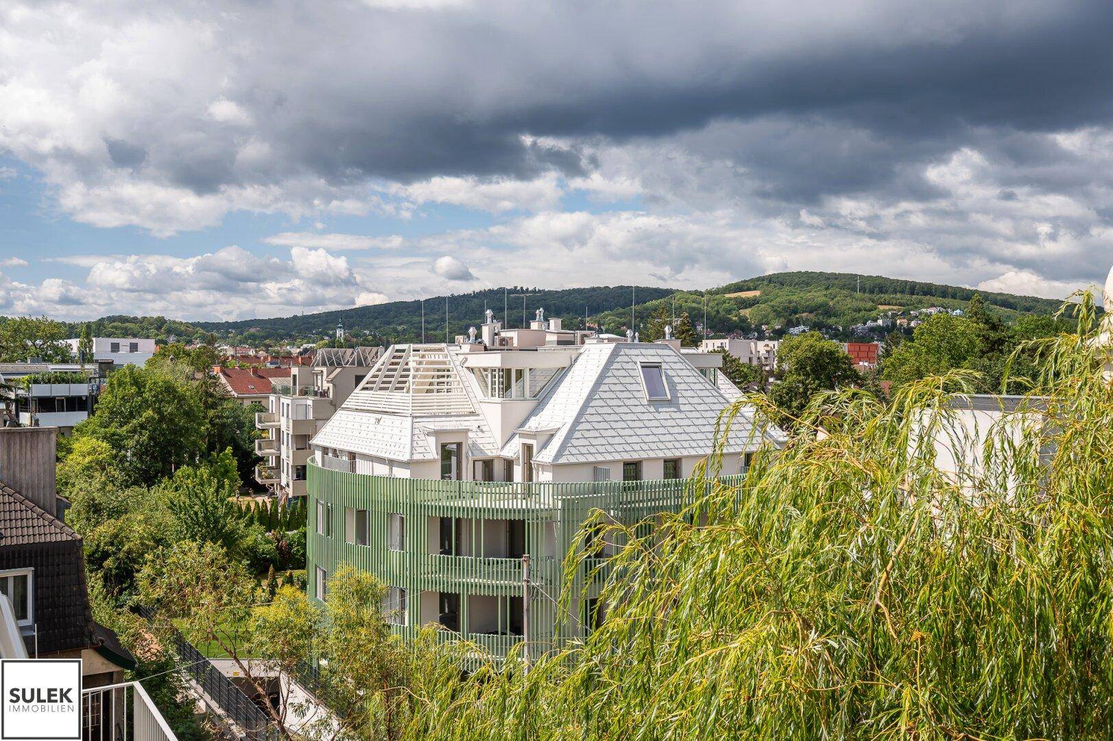 Modernes Gebäude mit grüner Lamellenfassade und Satteldach, umgeben von Bäumen und Hügeln.