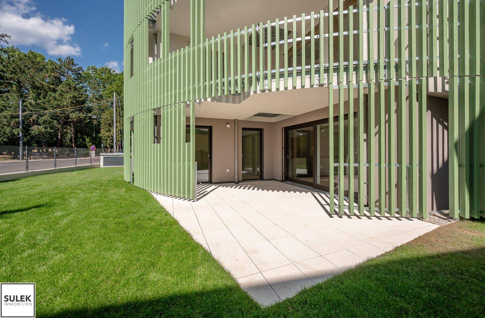 Außenansicht des Gebäudes mit grüner Lamellenfassade, Terrasse und gepflegtem Rasenbereich.