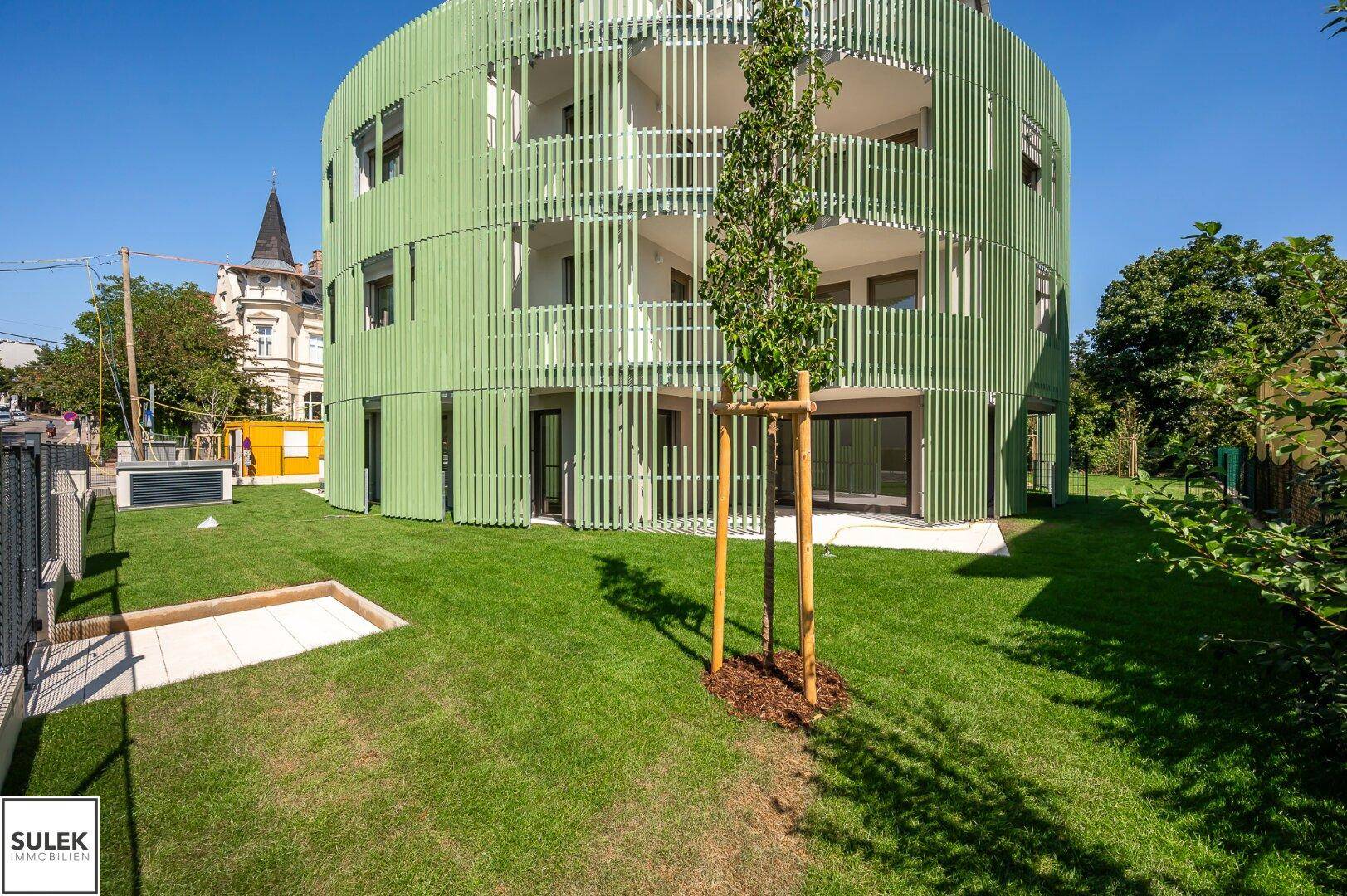 Modernes, rundes Gebäude mit grüner Lamellenfassade und gepflegter Rasenfläche im Vordergrund.