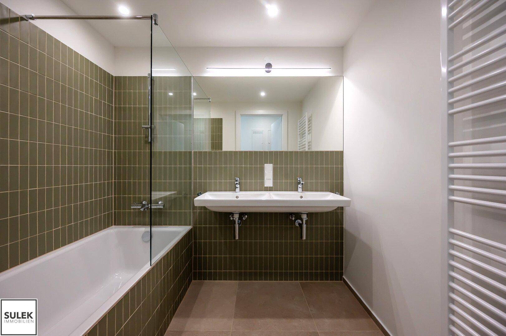 Modernes Badezimmer mit olivgrünen Fliesen, Badewanne, Doppelwaschbecken und Handtuchheizkörper.