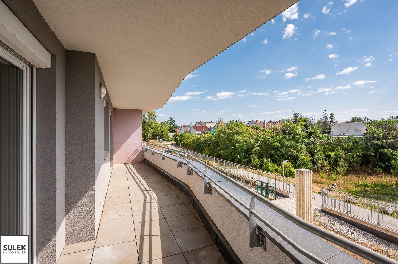 Langer Balkon mit Metallgeländer und Blick auf grüne Umgebung und Häuser.