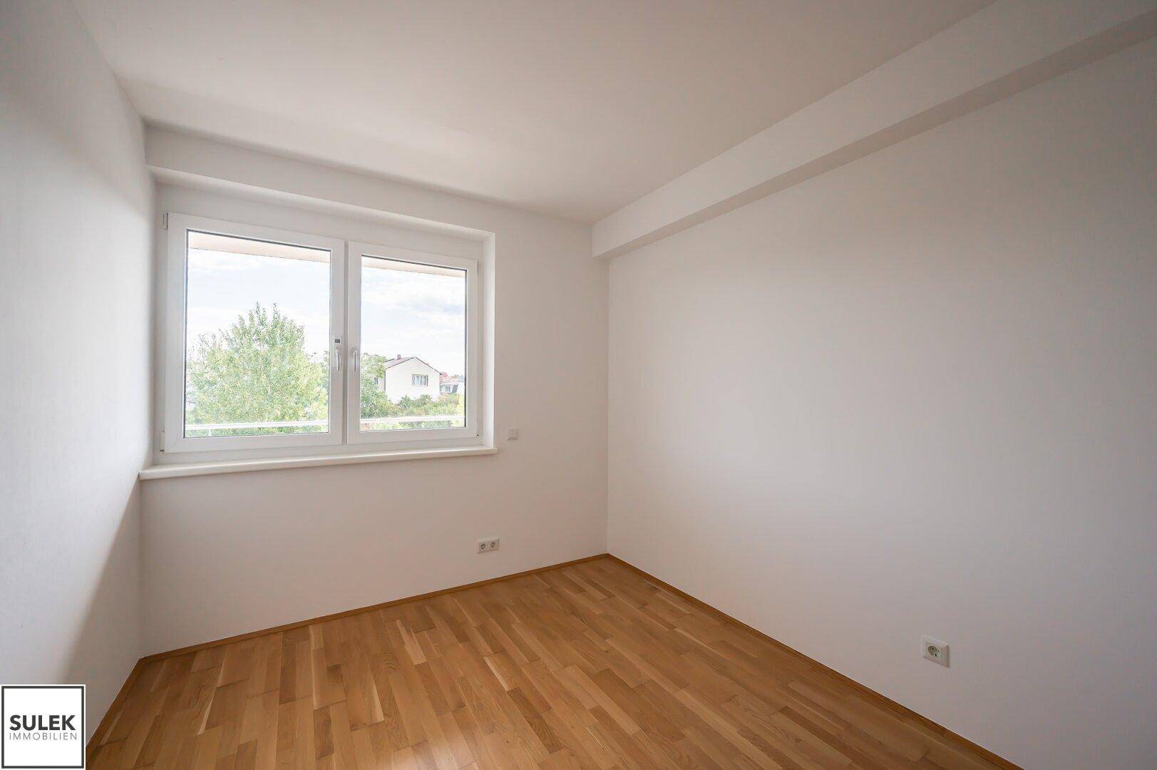 Helles, leeres Zimmer mit Holzparkettboden und großem Fenster mit Blick ins Grüne.