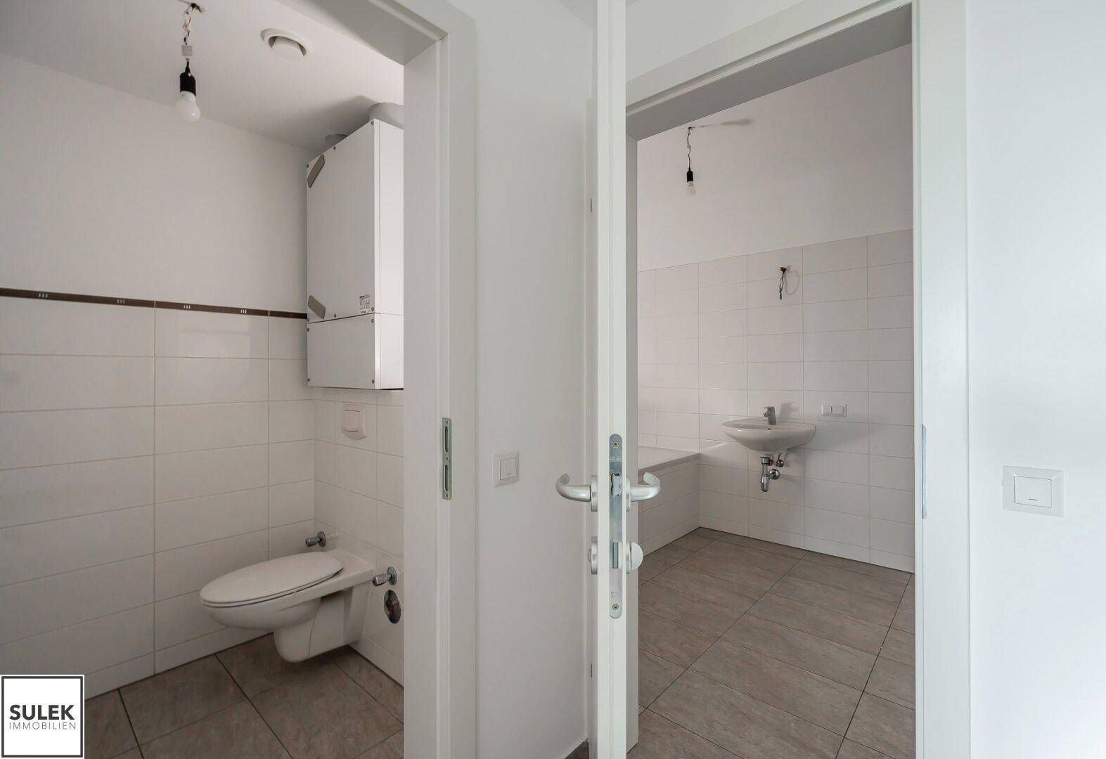 Separates WC mit Wand-WC und kleinem Waschbecken, komplett gefliest in Weiß.