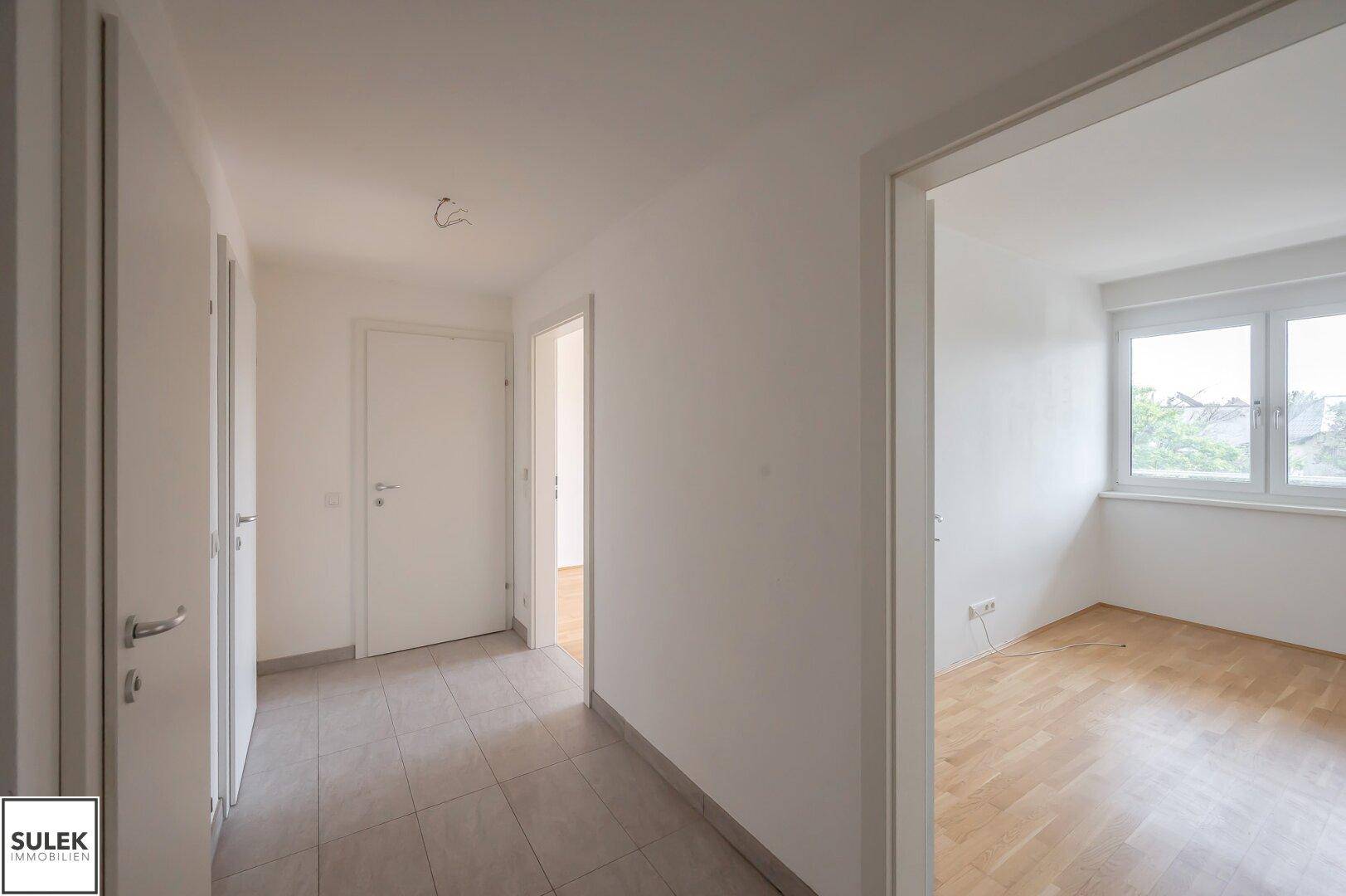 Flur mit Fliesenboden, der zu einem hellen Zimmer mit Parkett und Fenster führt.
