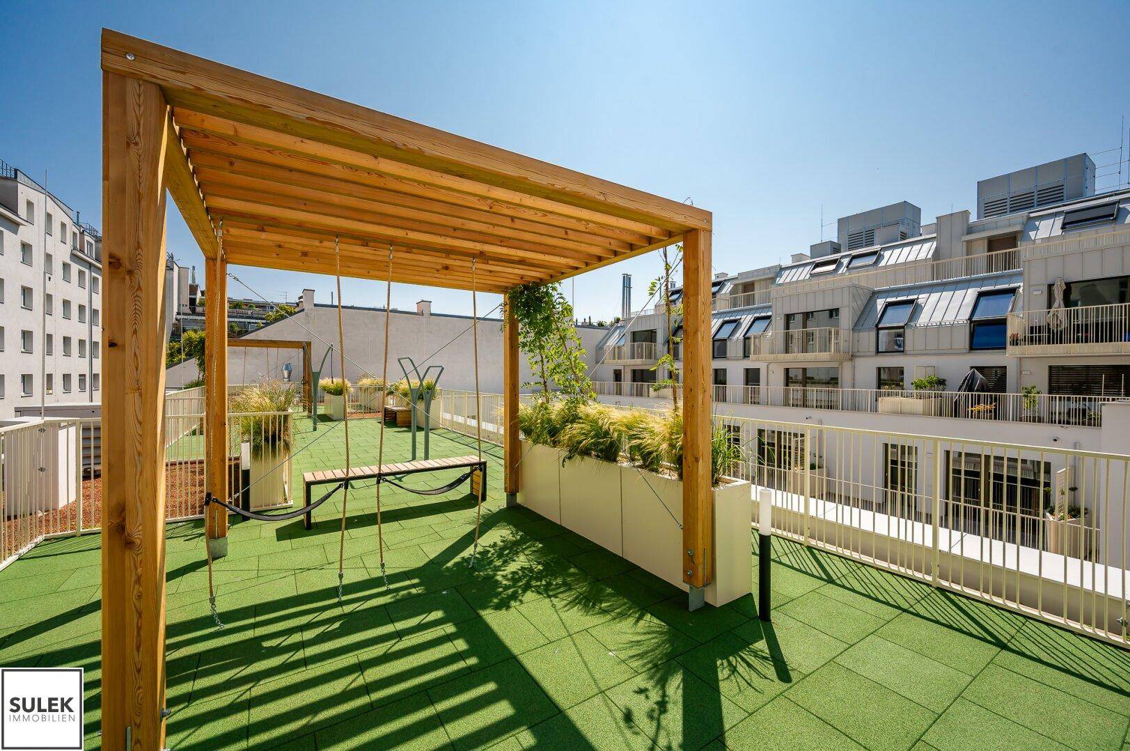 Sonnige Dachterrasse mit grüner Bodenfläche, Holzpergola und gemütlicher Schaukelbank, umgeben von modernen Gebäuden.