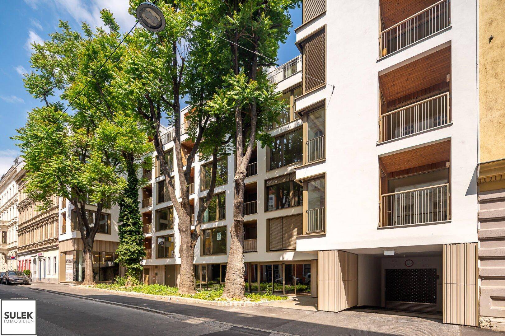 Moderne Wohnfassade mit großen Fenstern und Balkonen, umgeben von hohen Bäumen an einer Stadtstraße.
