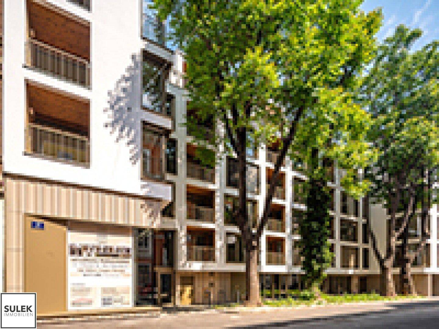 Ansicht einer modernen Wohnanlage mit großen Fenstern und Balkonen, gesäumt von Bäumen an der Straße.