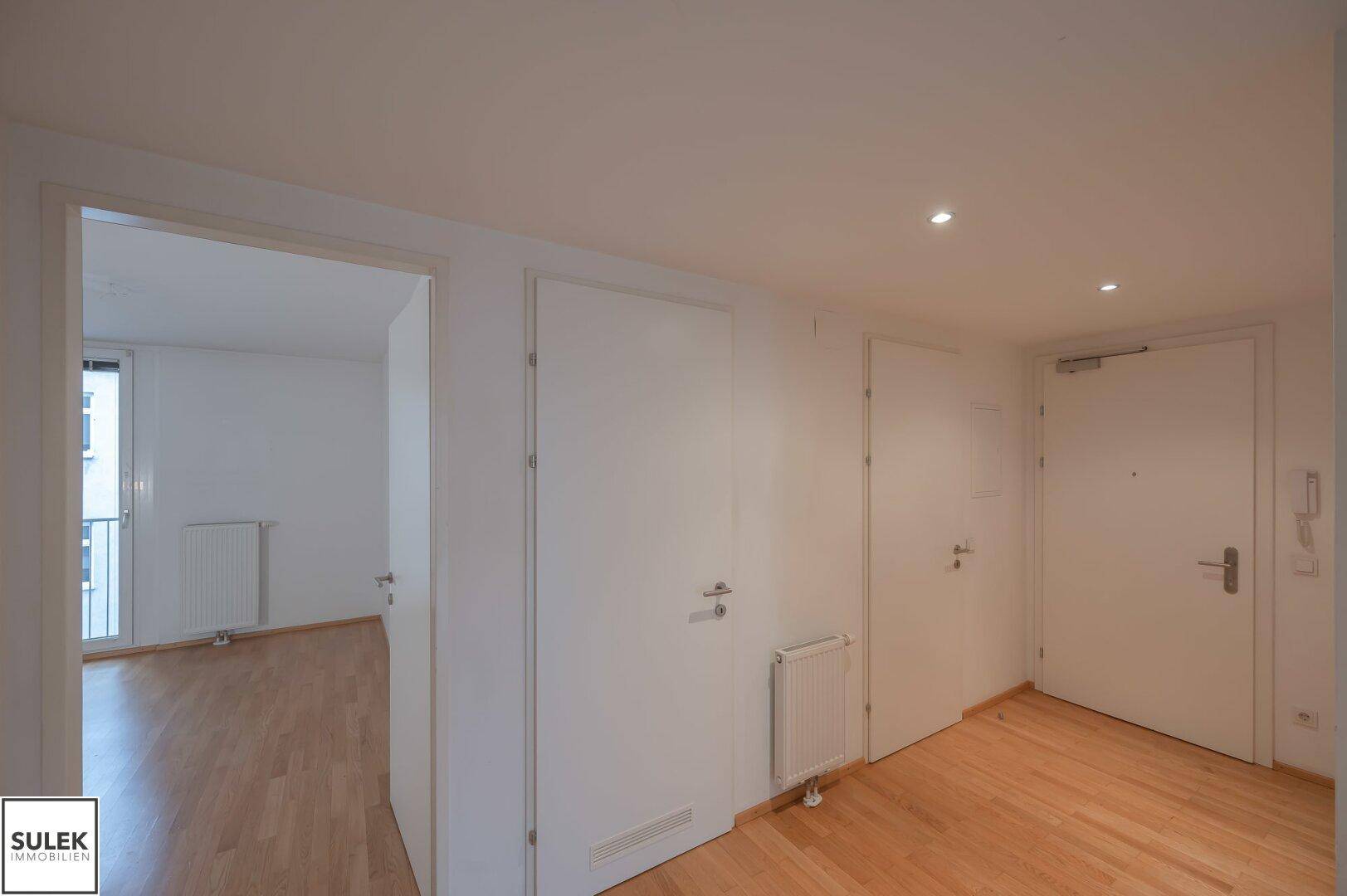 Langer Flur mit mehreren Türen und Blick in ein Zimmer mit Balkonzugang.