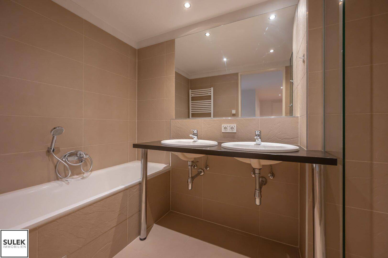 Modernes Badezimmer mit Badewanne, Doppelwaschbecken und großem Spiegel, hell gefliest.