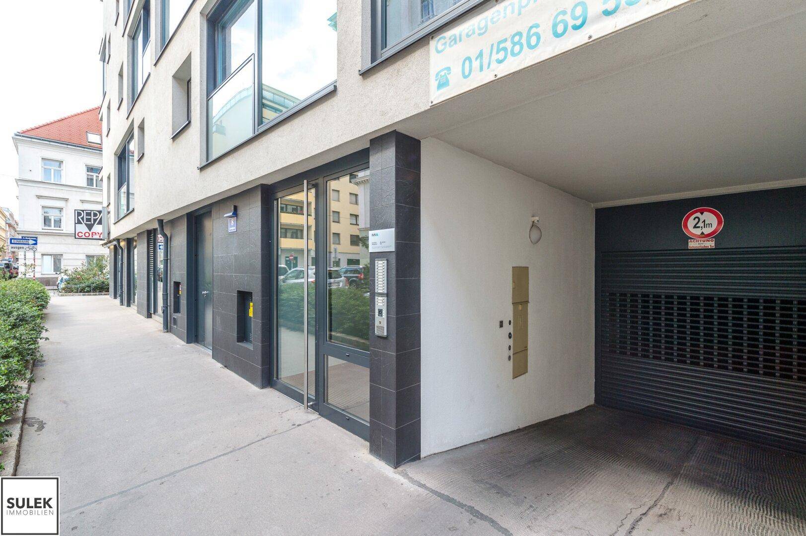 Gebäudeeingang mit moderner Fassade und Zufahrt zu einer Tiefgarage an der Straße.