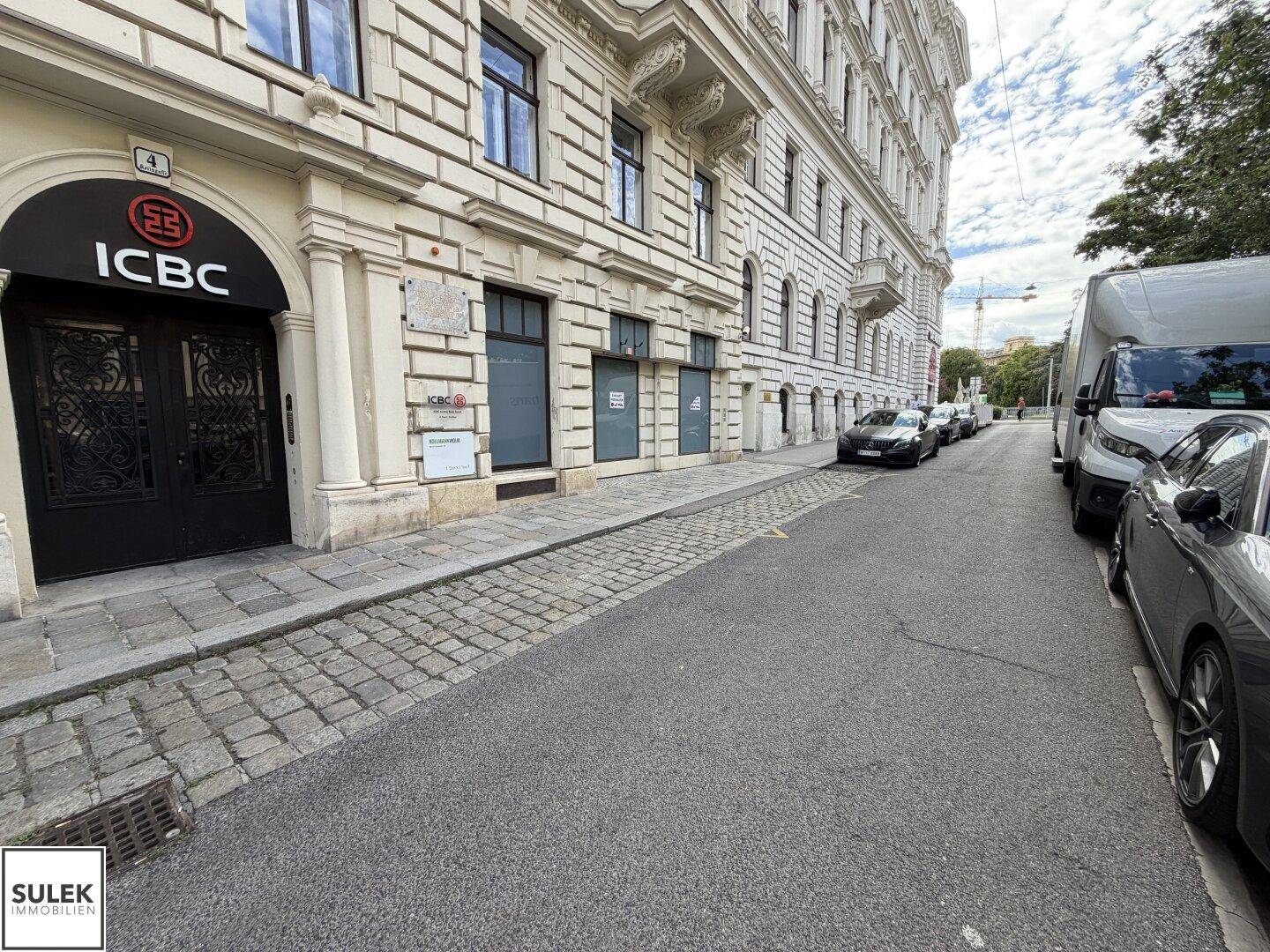 Straßenansicht eines Gebäudes mit klassischer Architektur, Bankfiliale und geparkten Fahrzeugen.