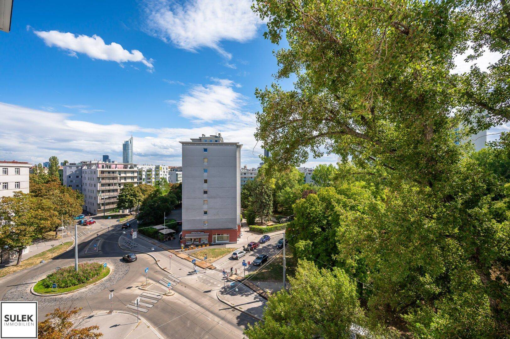 Panoramablick über eine belebte Stadtstraße mit Gebäuden, Bäumen und blauem Himmel.