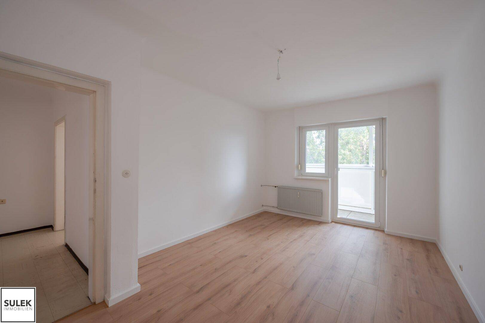 Geräumiges Zimmer mit hellem Laminatboden, großen Fenstern und Zugang zum Balkon.