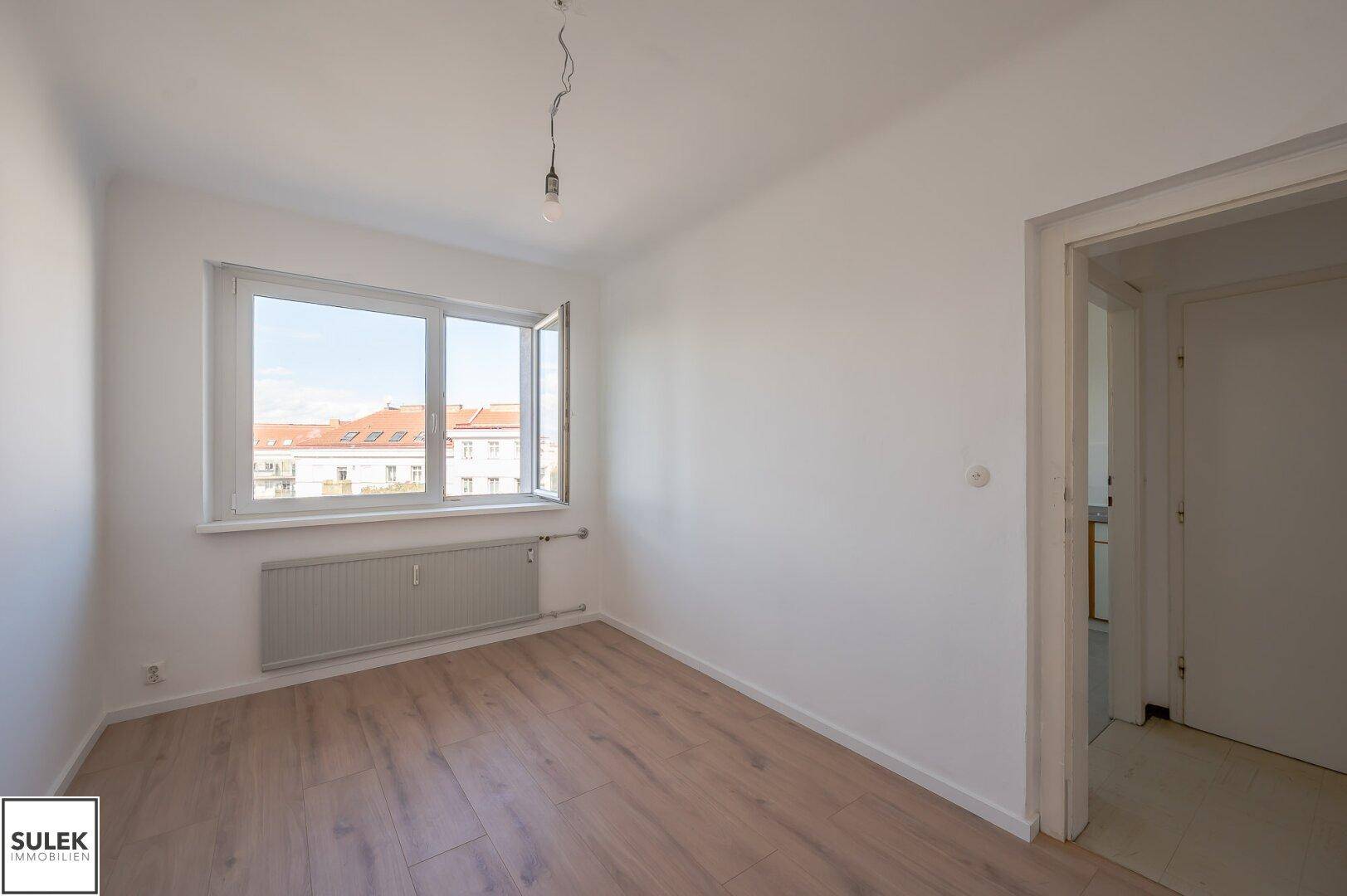 Freundliches Zimmer mit Laminatboden, weiß gestrichenen Wänden und Blick auf die Stadt.
