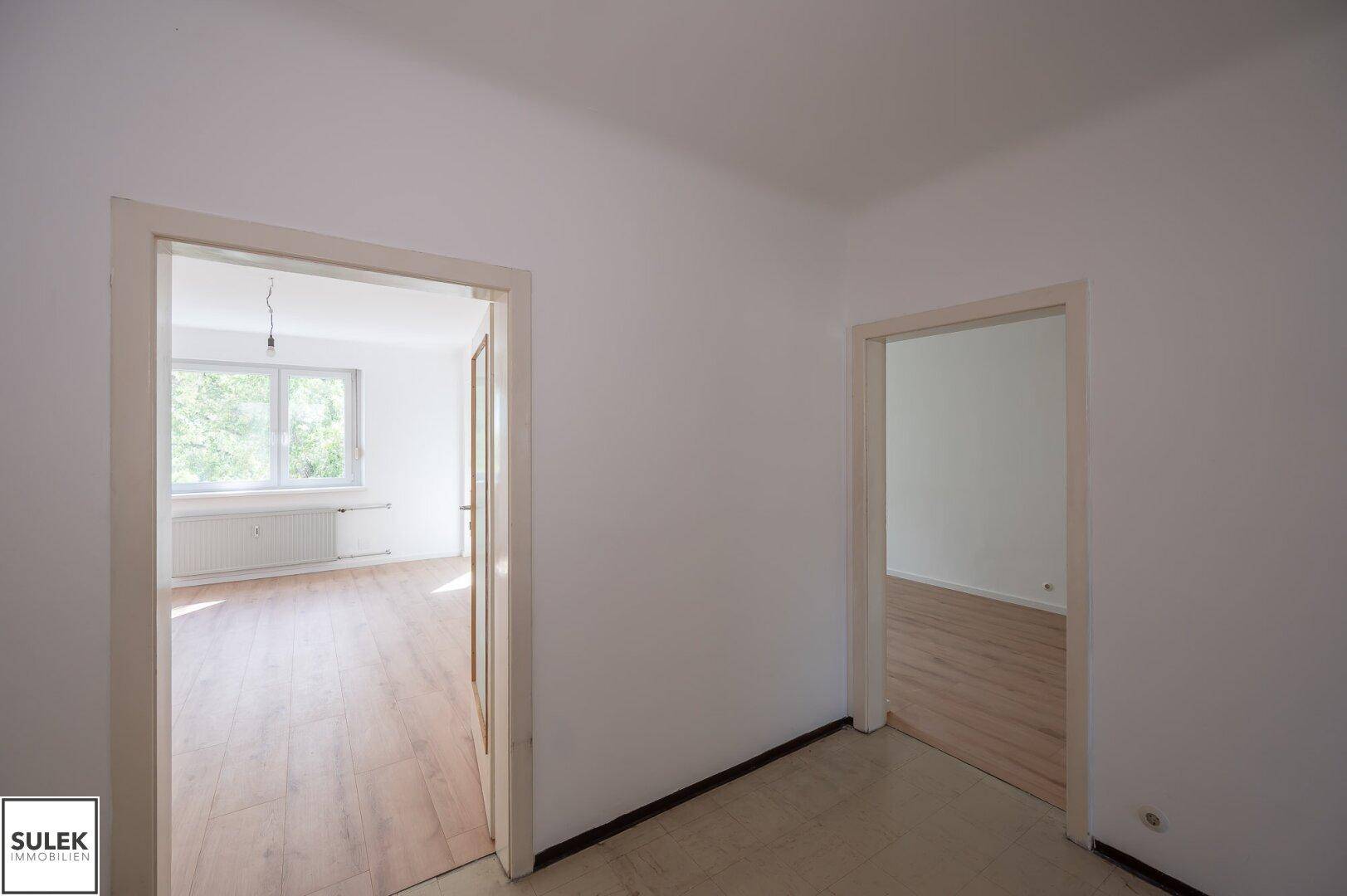Blick durch eine Tür in ein weiteres helles Zimmer mit Laminatboden und Fenster.