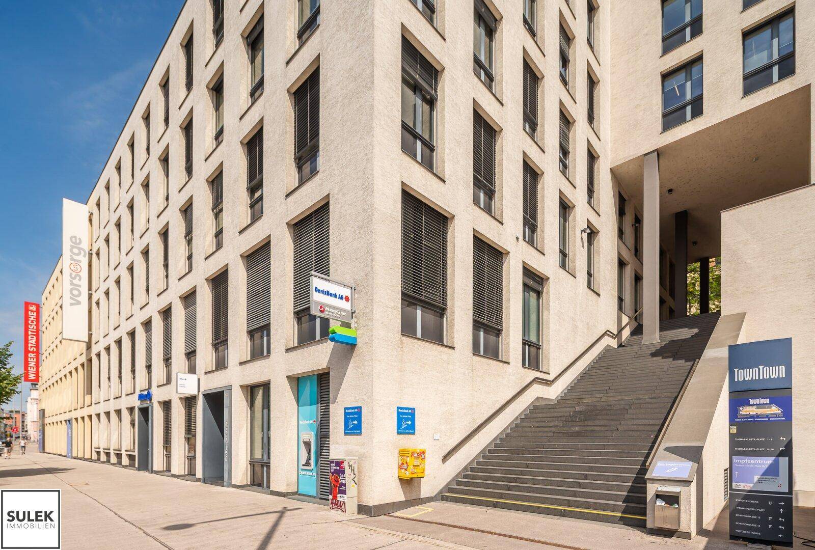 Außenansicht eines modernen Bürogebäudes mit Treppe und Geschäften im Erdgeschoss an einem sonnigen Tag.