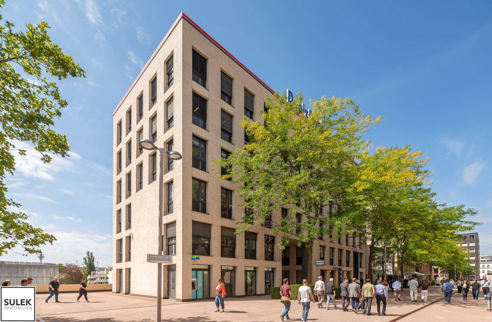 Gesamtansicht eines modernen Bürogebäudes mit mehreren Etagen und Bäumen davor an einem sonnigen Tag.