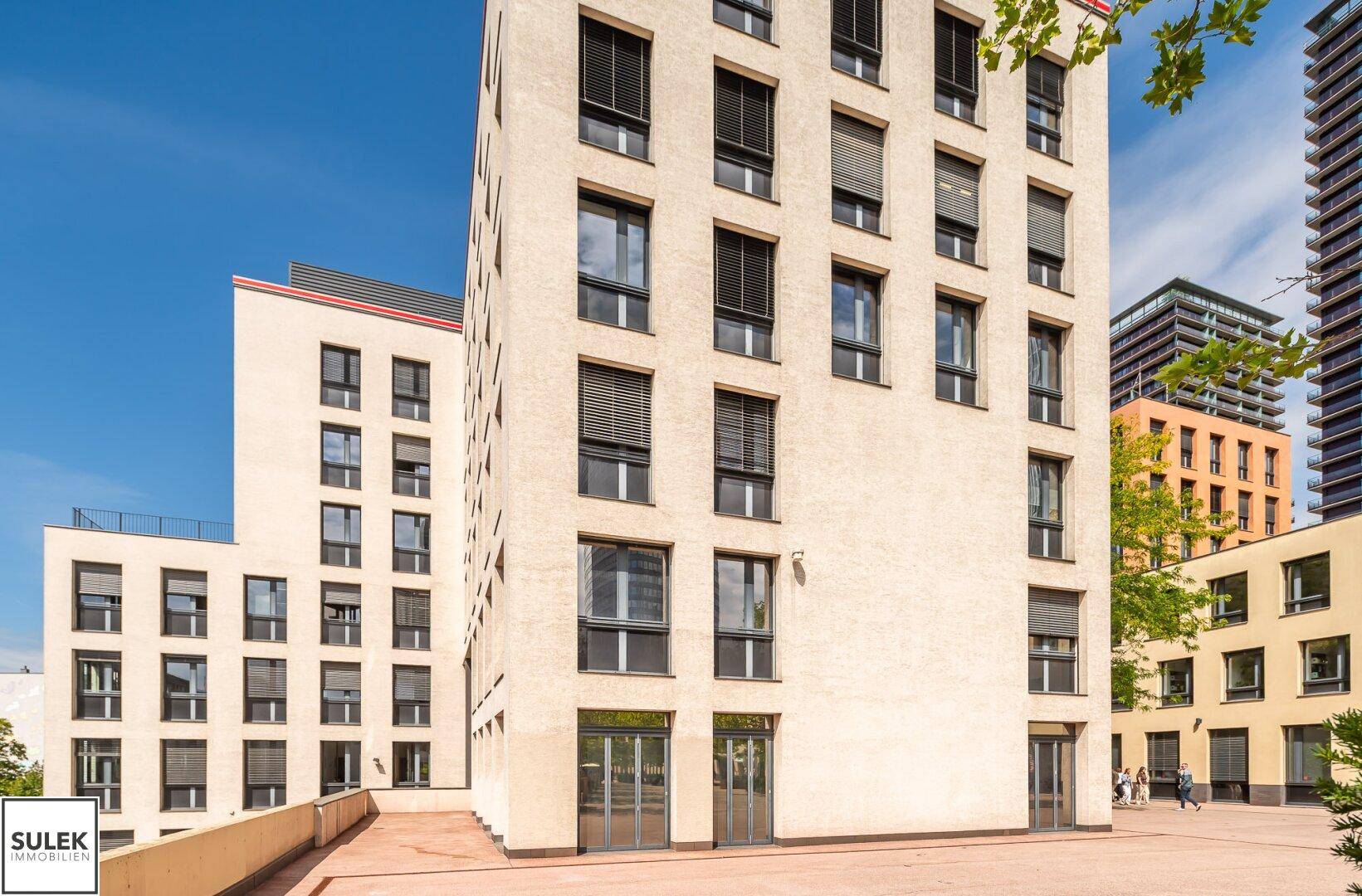 Seitenansicht eines modernen Bürogebäudes mit vielen Fenstern und angrenzenden Gebäuden unter blauem Himmel.