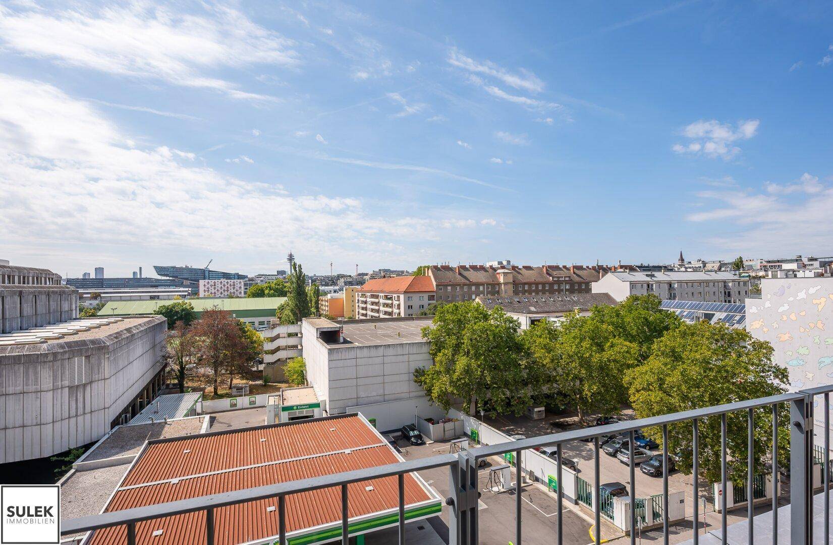 Balkon mit Metallgeländer bietet einen weiten Blick über die Stadt und grüne Bäume unter blauem Himmel.