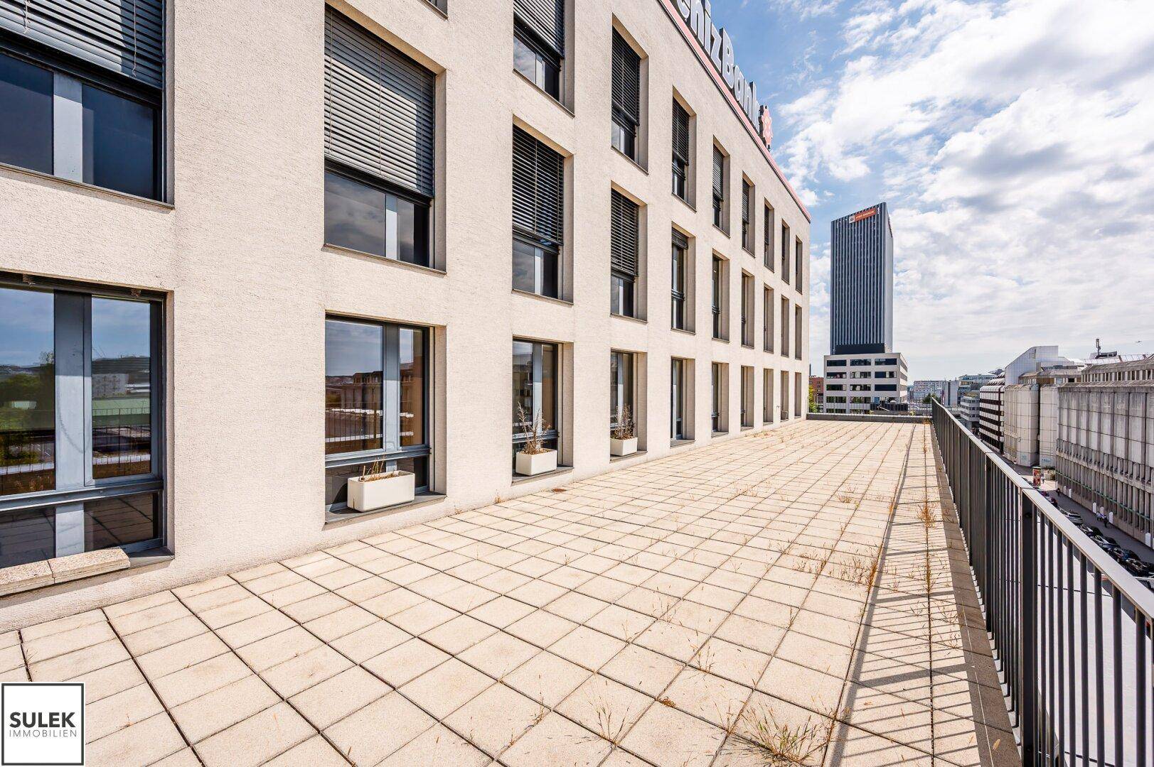 Weitläufige Terrasse mit Fliesenboden entlang der Gebäudefassade und Blick auf die Stadt.