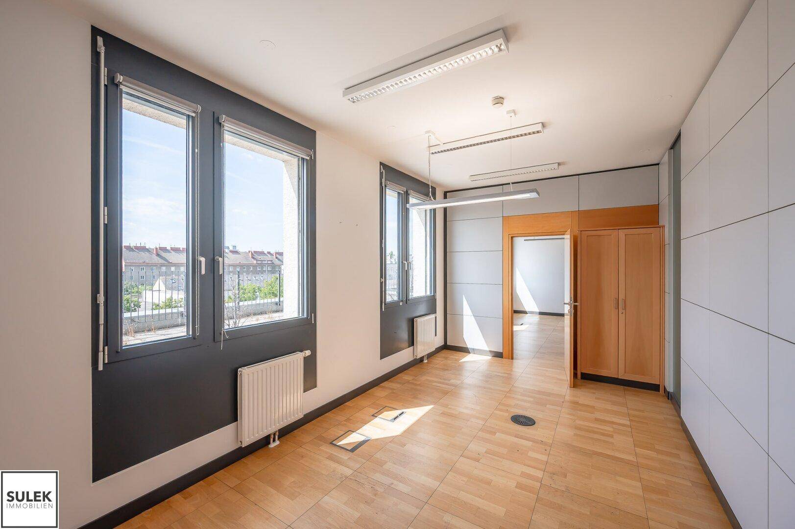 Helles Büro mit Holzparkett, großen Fenstern und Blick auf die Stadt unter blauem Himmel.