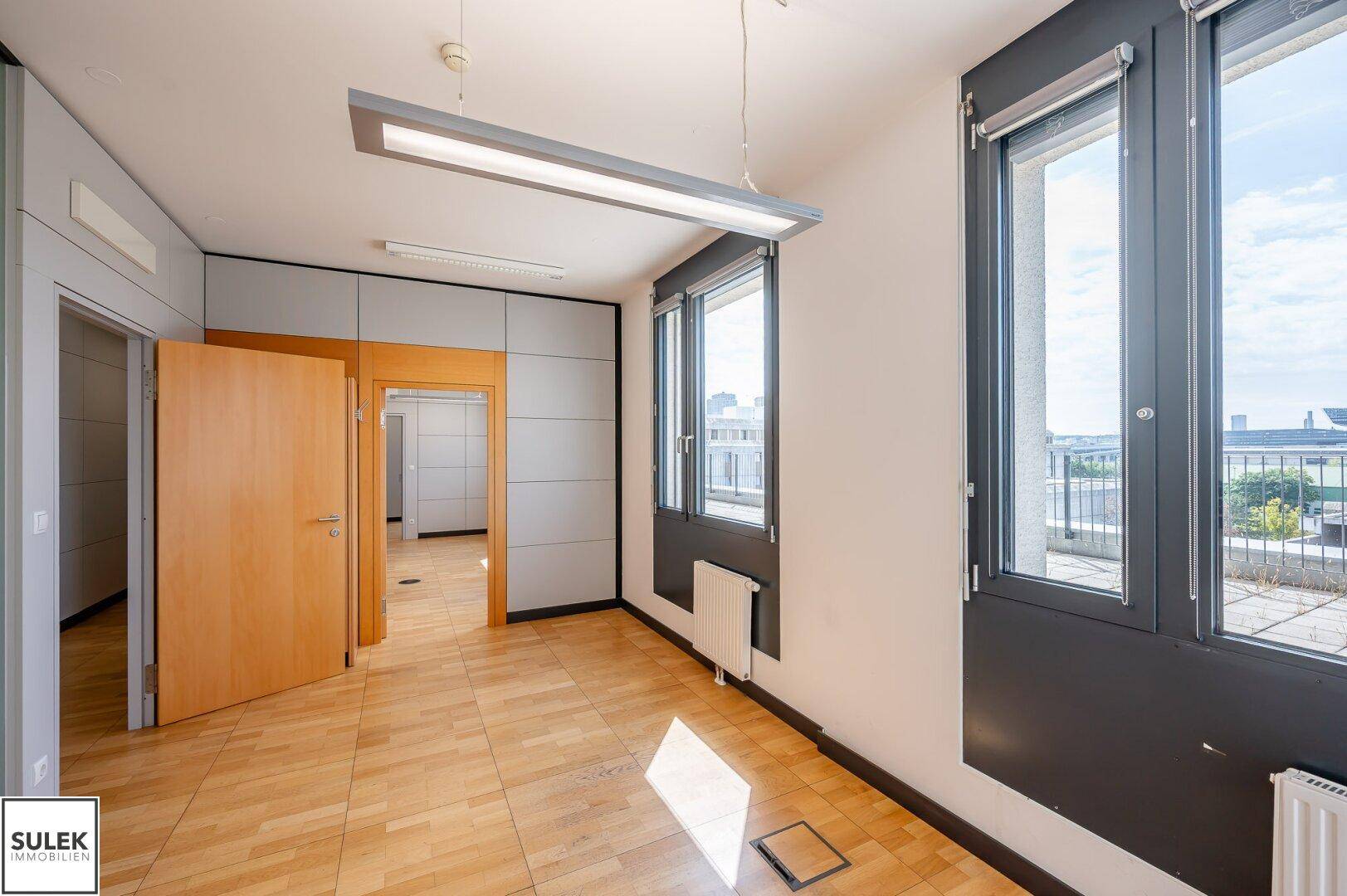 Geräumiges Büro mit Holzboden, großen Fenstern und Zugang zu weiteren Räumen in einem Bürogebäude.
