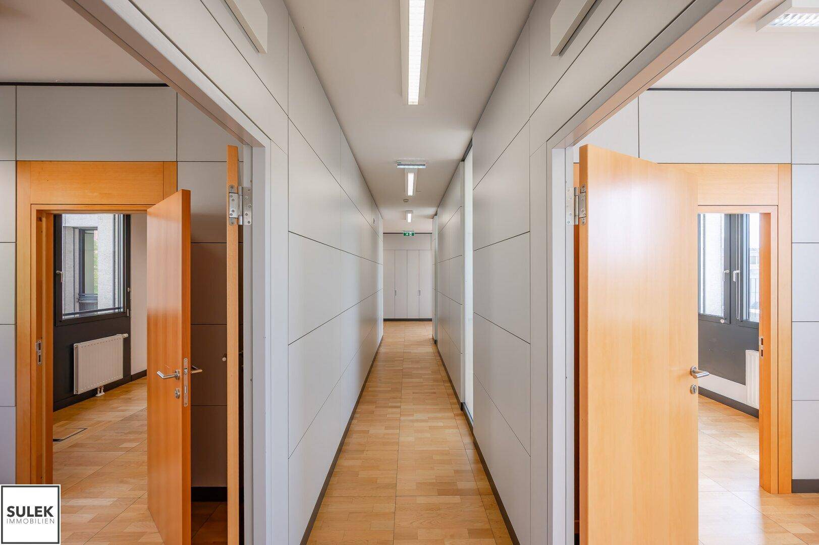 Langer Flur mit Holzboden, hellen Wänden und mehreren Türen zu Büroräumen.