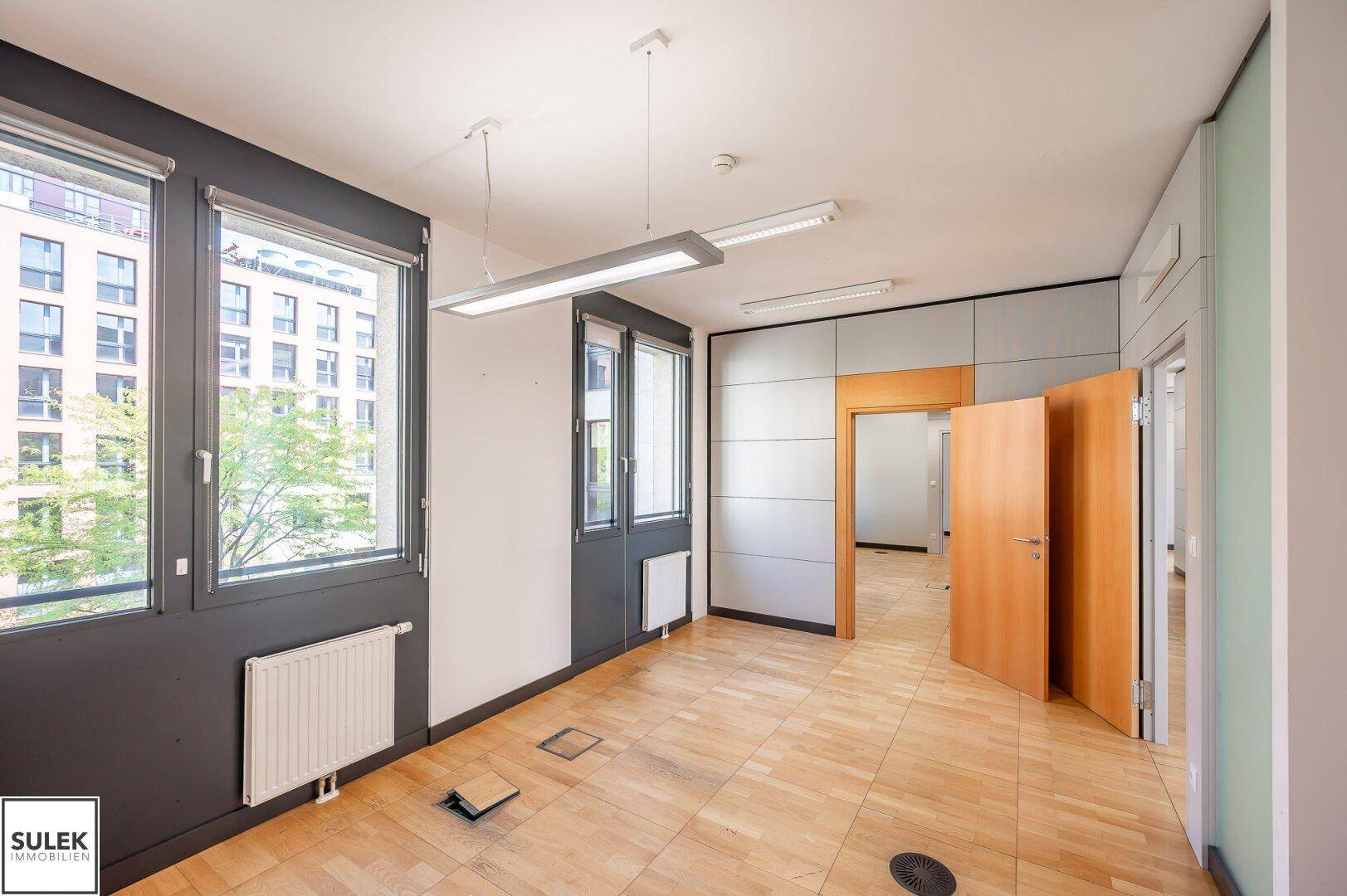 Helles Büro mit Holzboden, großen Fenstern und Blick auf die grüne Umgebung.
