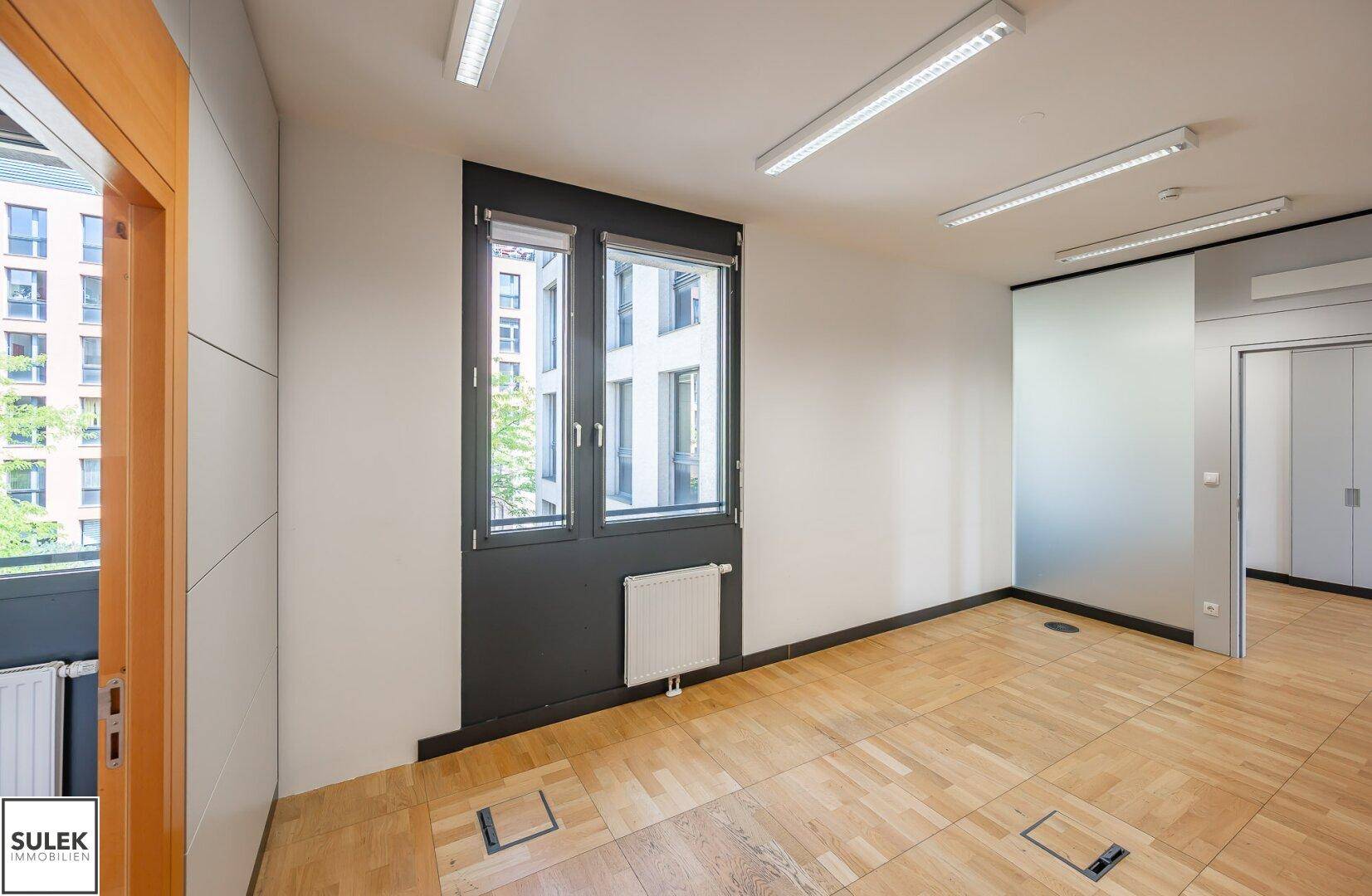 Büro mit Holzboden, großen Fenstern und Blick auf die umliegenden Gebäude.
