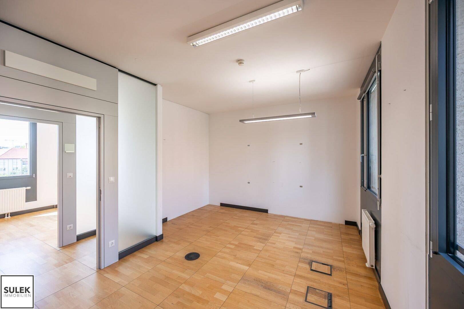 Leeres Büro mit Holzboden, weißen Wänden und Blick auf die Umgebung durch Fenster.