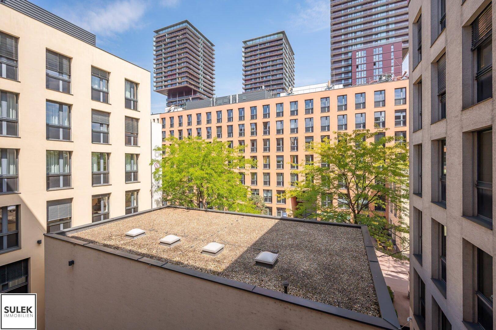 Blick auf die Dächer umliegender Gebäude und moderne Hochhäuser unter blauem Himmel.