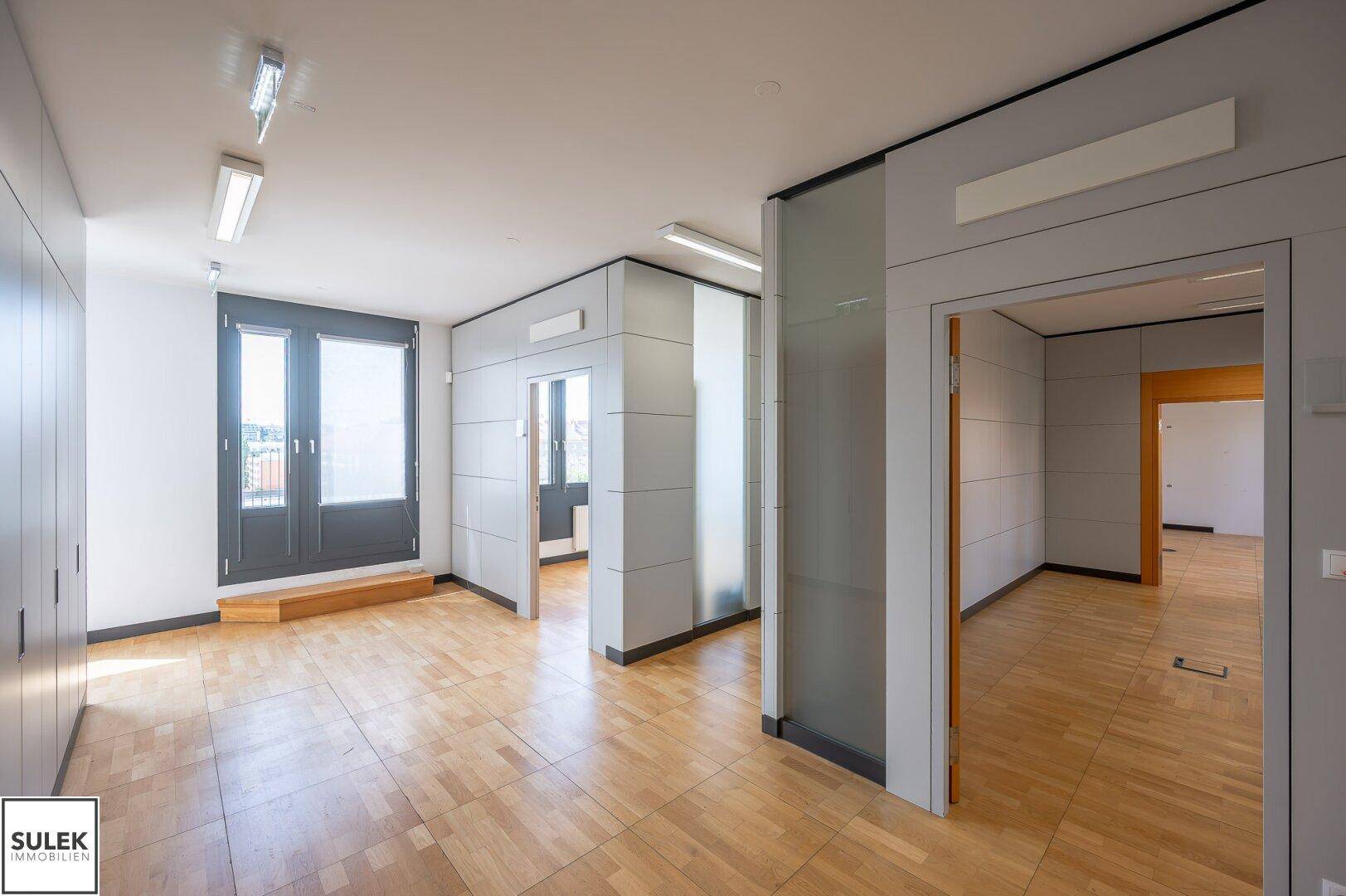 Büro mit Holzboden, großen Fenstern und Zugang zu weiteren Räumen.