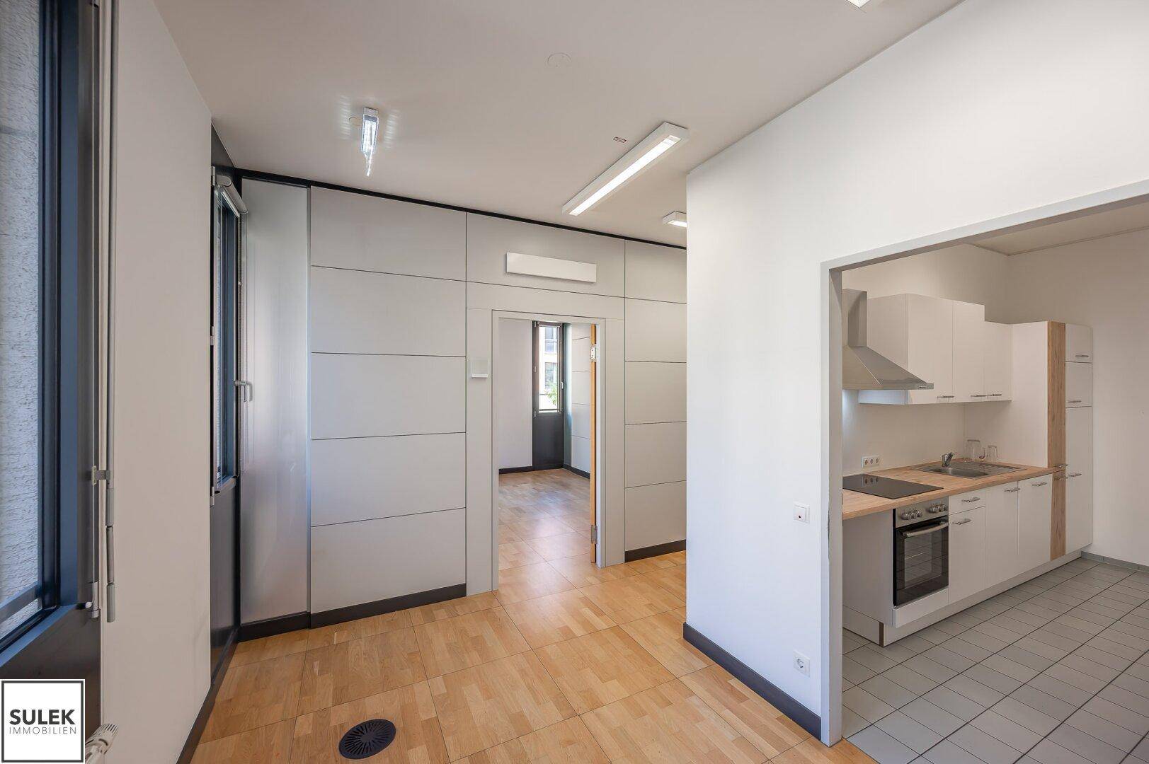 Büro mit Holzboden und angrenzender moderner Küchenzeile mit weißen Schränken.
