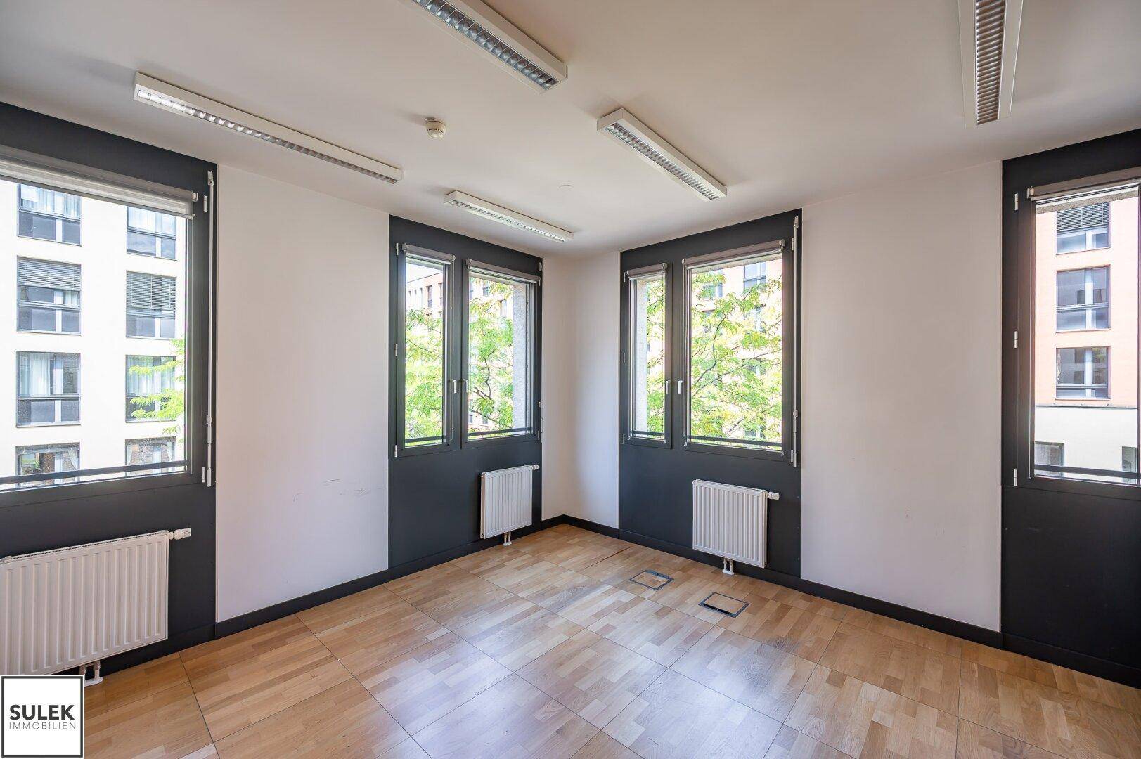 Heller Büroraum mit Parkettboden, großen Fenstern und Blick auf umliegende Gebäude.