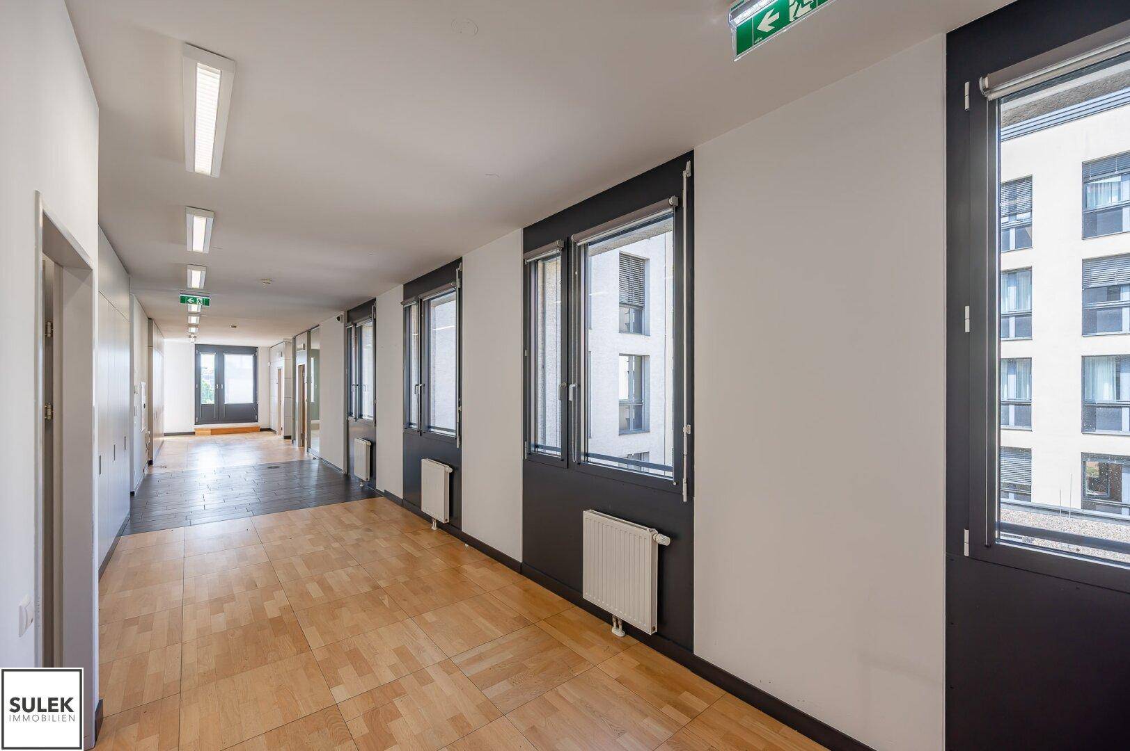 Langer Flur mit Holzboden, Fenstern und Blick auf die umliegenden Gebäude.