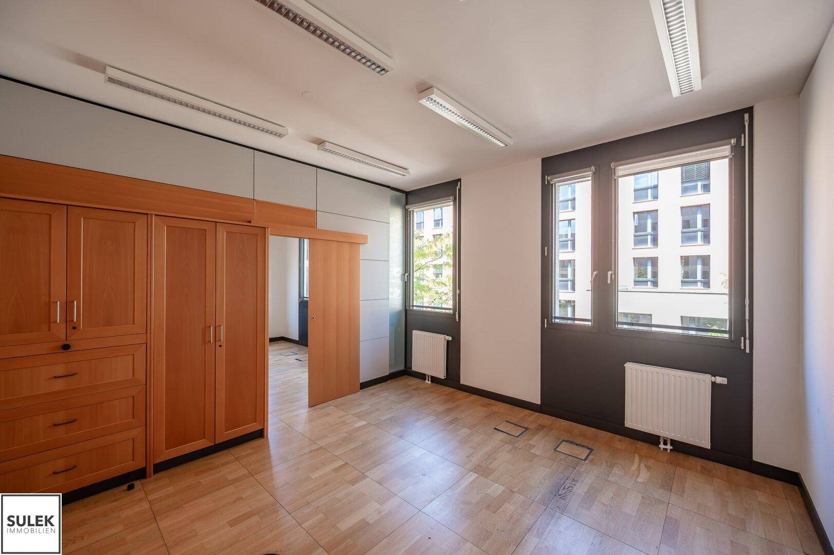 Büroraum mit Holzboden, Einbauschrank und Blick durch die Fenster auf andere Gebäude.