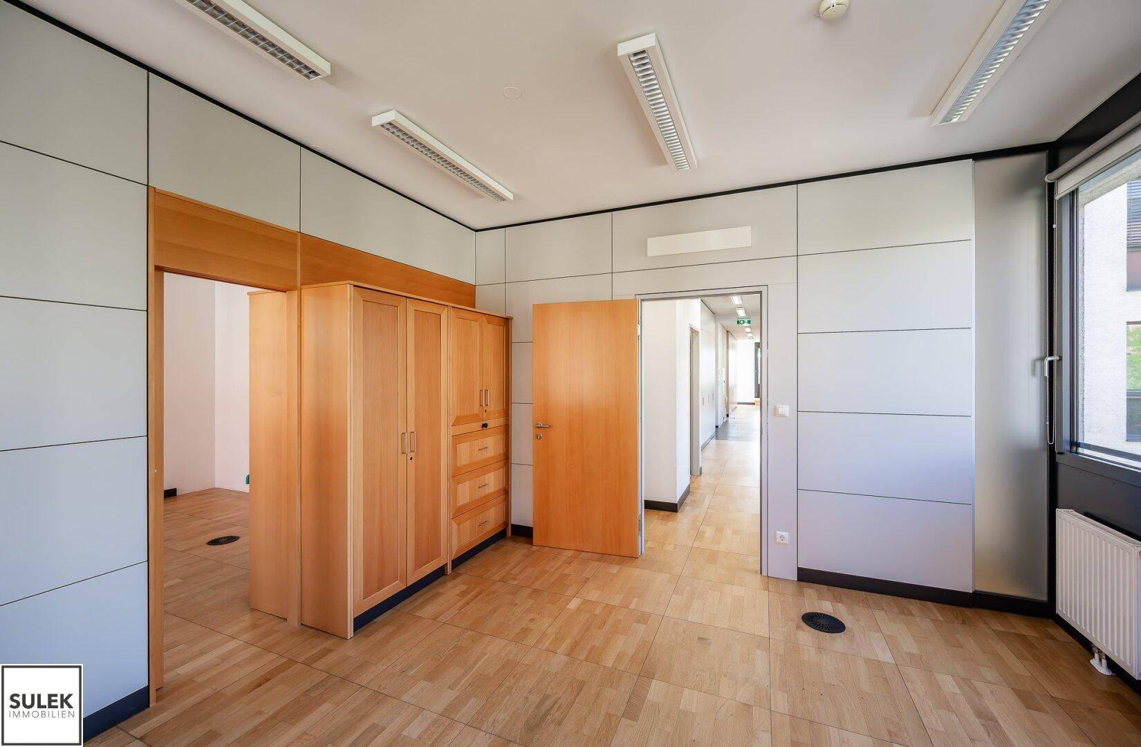 Büroraum mit Holzboden, Einbauschrank und Zugang zu einem Flur.