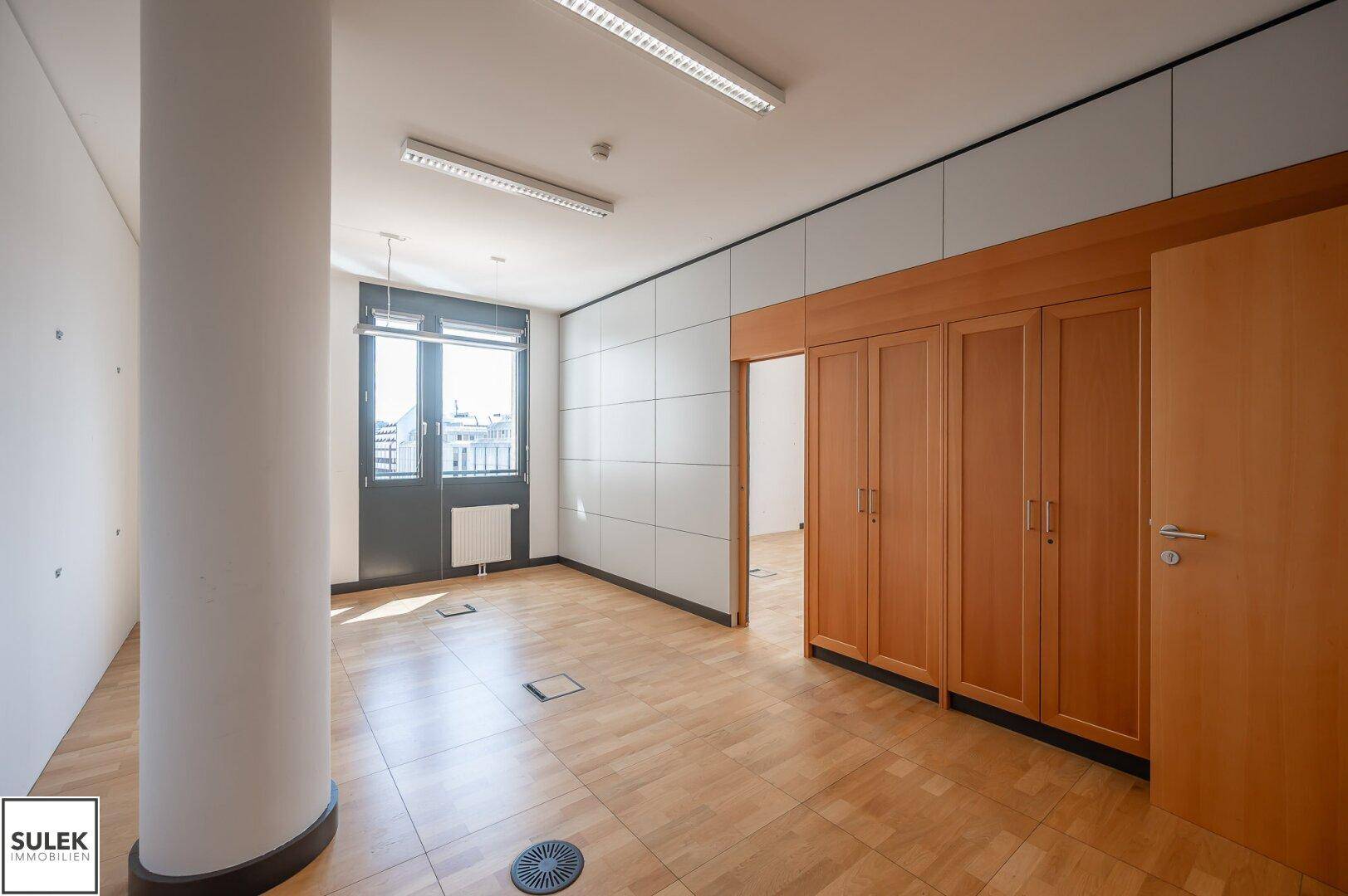 Büroraum mit Holzboden, runder Säule und Einbauschränken entlang der Wand.
